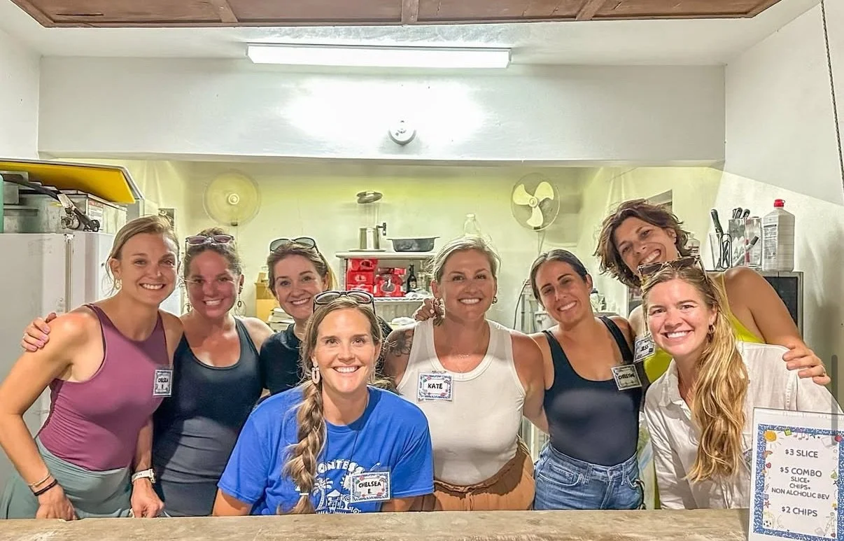 Group of women smiling in a kitchen, some wearing casual clothing, standing together behind a counter with a menu sign on the right side listing food prices.