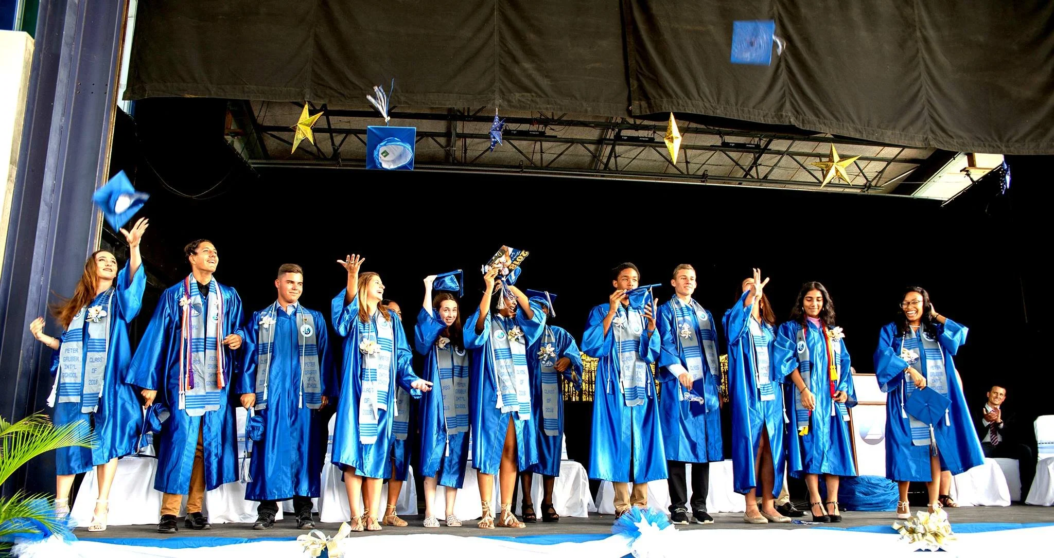Graduates in blue gowns and stoles celebrating on stage at a graduation ceremony, some tossing caps in the air.