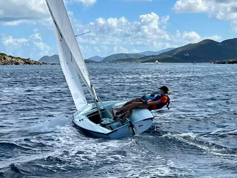 Person sailing a small boat or dinghy on open water, with mountains and cloudy sky in the background.