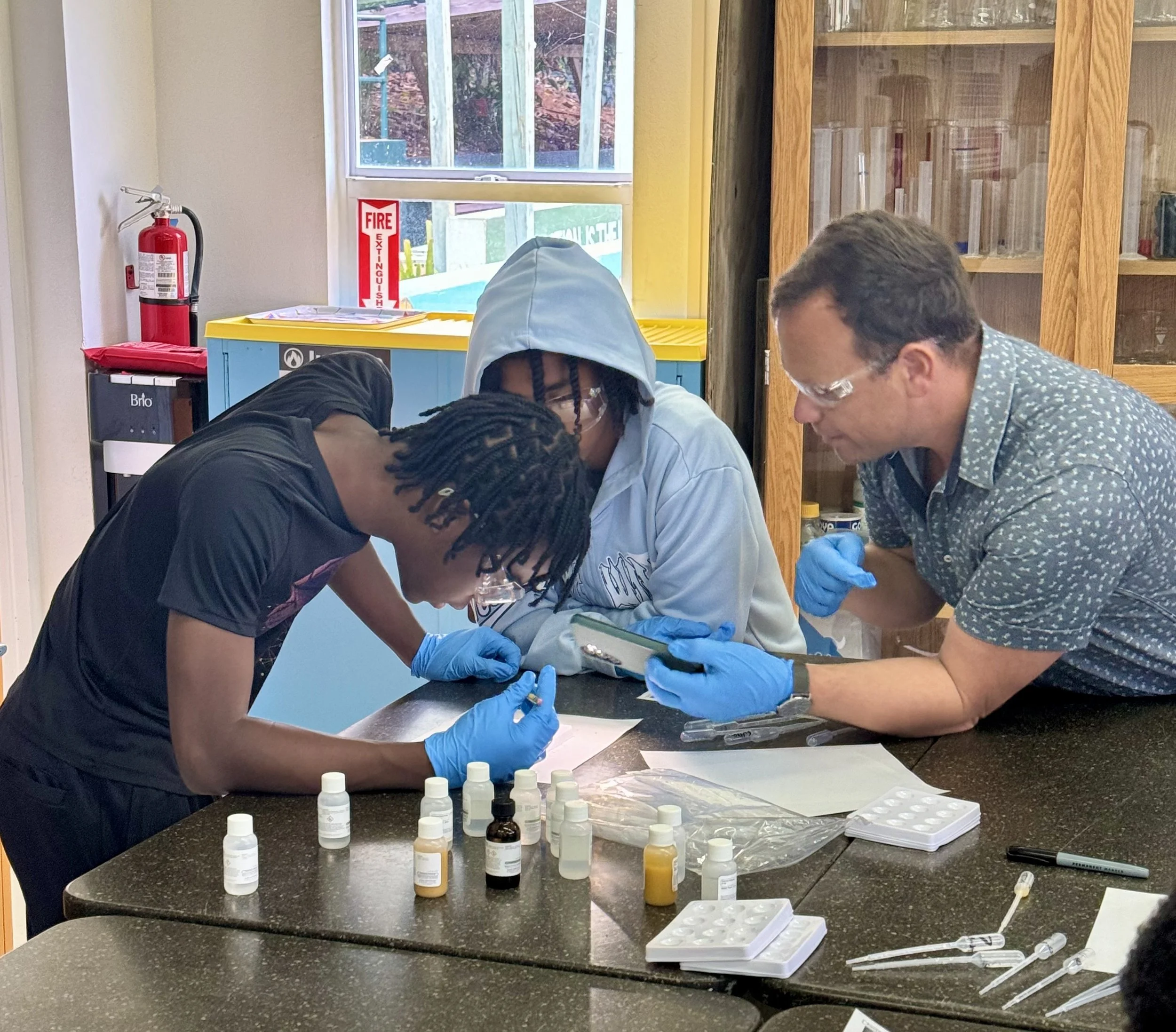 Three people wearing safety glasses and blue gloves are working together on a scientific experiment at a table with bottles, pipettes, and lab equipment, inside a room with a fire extinguisher and a window.