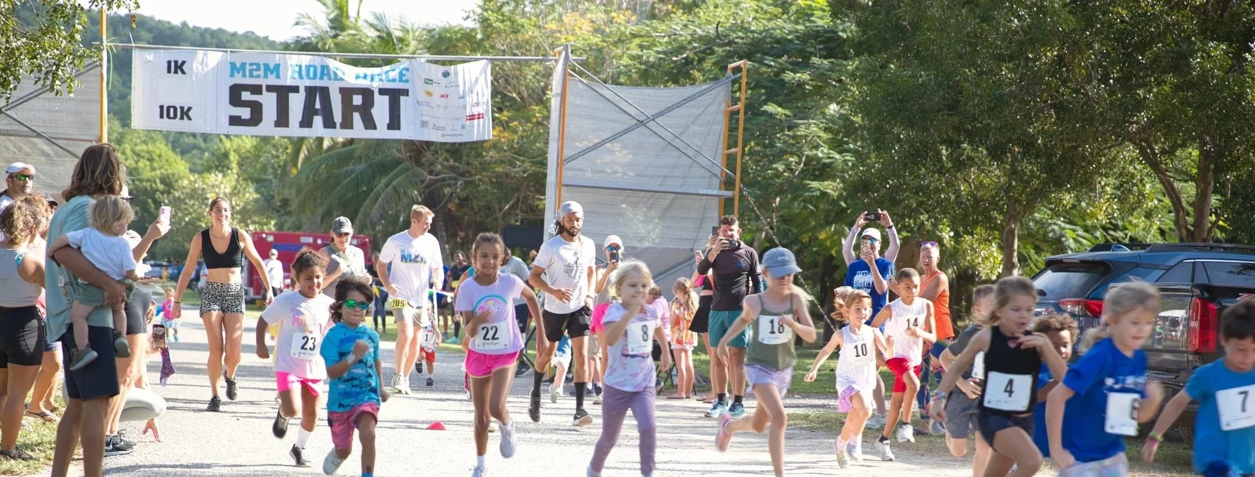 Children and adults participating in a 1K or 10K race start event, with some spectators taking photos, under a start banner on a sunny day with green trees in the background.