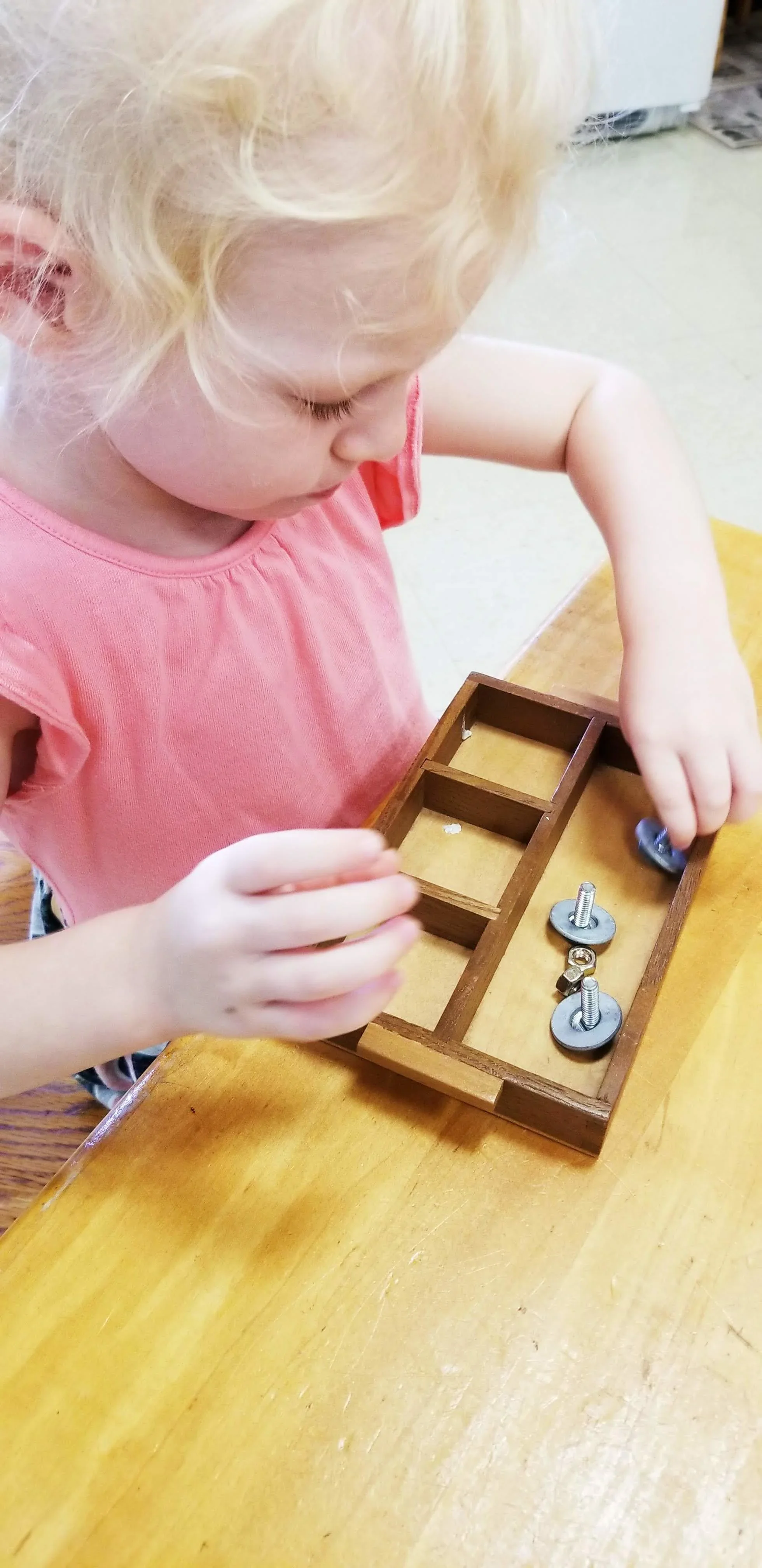 A young girl with blonde curly hair working on a small wooden project involving wheels and bolts, seated at a wooden table.
