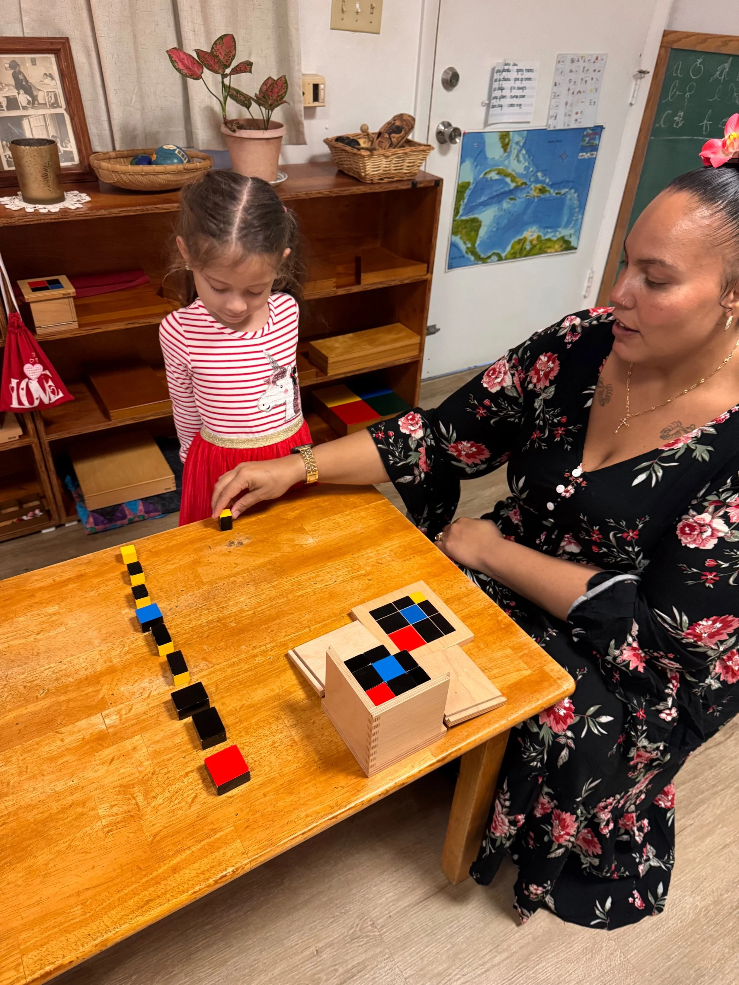 A young girl in a red and white striped shirt with a unicorn on it, and a woman in a black floral dress, play a game with black, yellow, red, and blue tiles on a wooden table in a classroom setting.