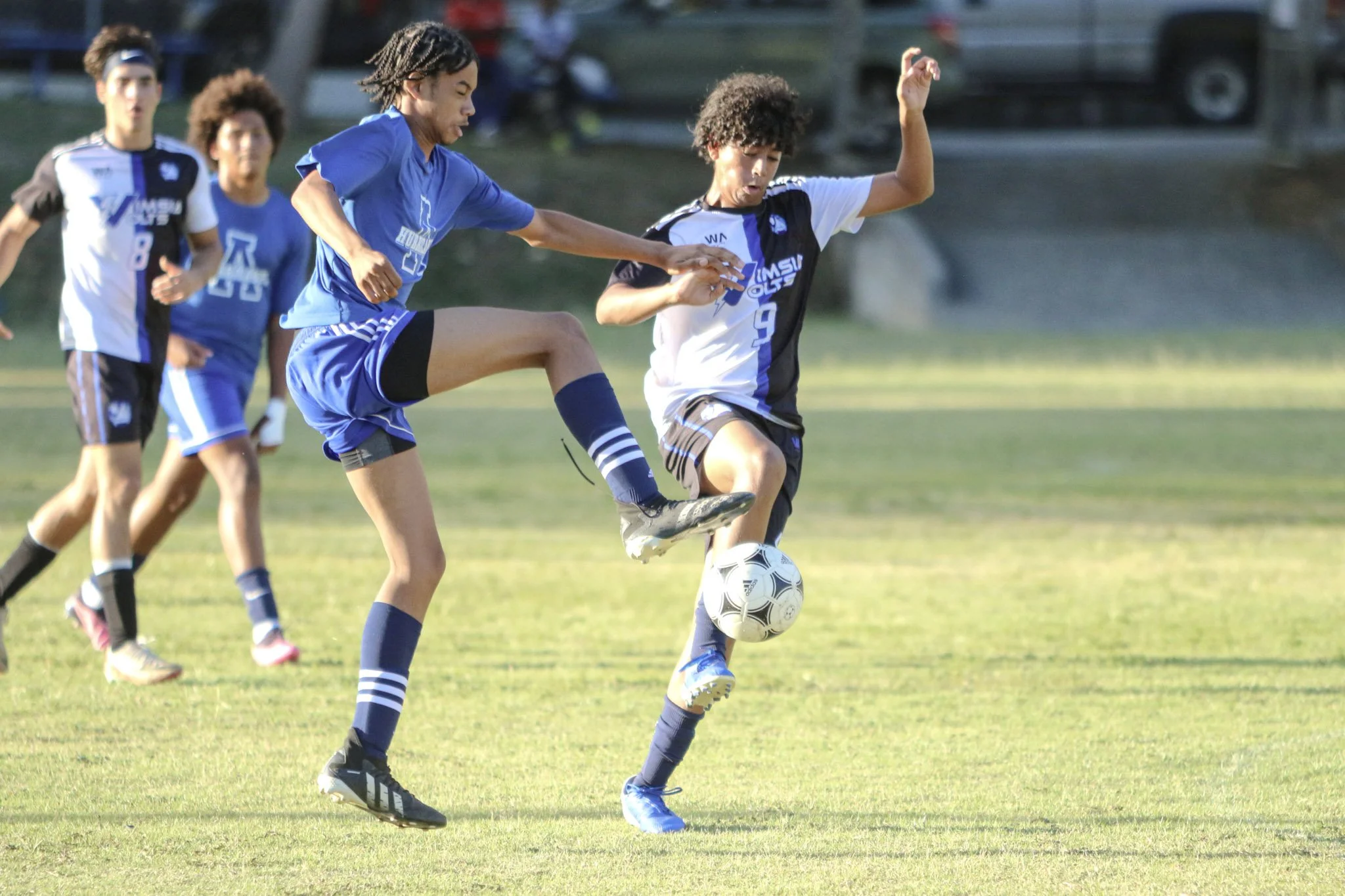 Young boys playing soccer on a grassy field, with two players actively contesting the ball while others watch in the background.