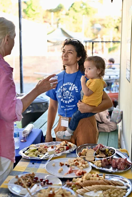 A woman holding a young child at a table with various food platters, engaging in conversation with an elderly woman at an outdoor gathering.