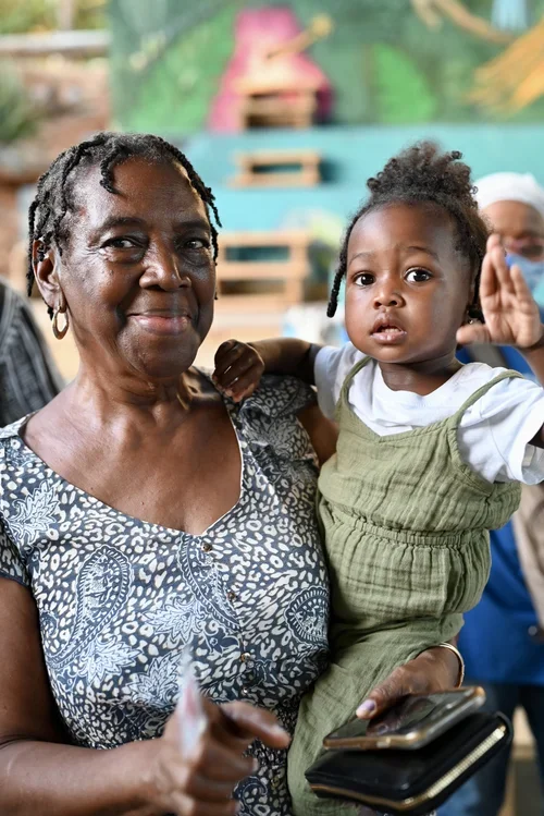 An elderly woman holding a young child with curly hair, wearing a green outfit, in a vibrant indoor setting with a colorful mural background.