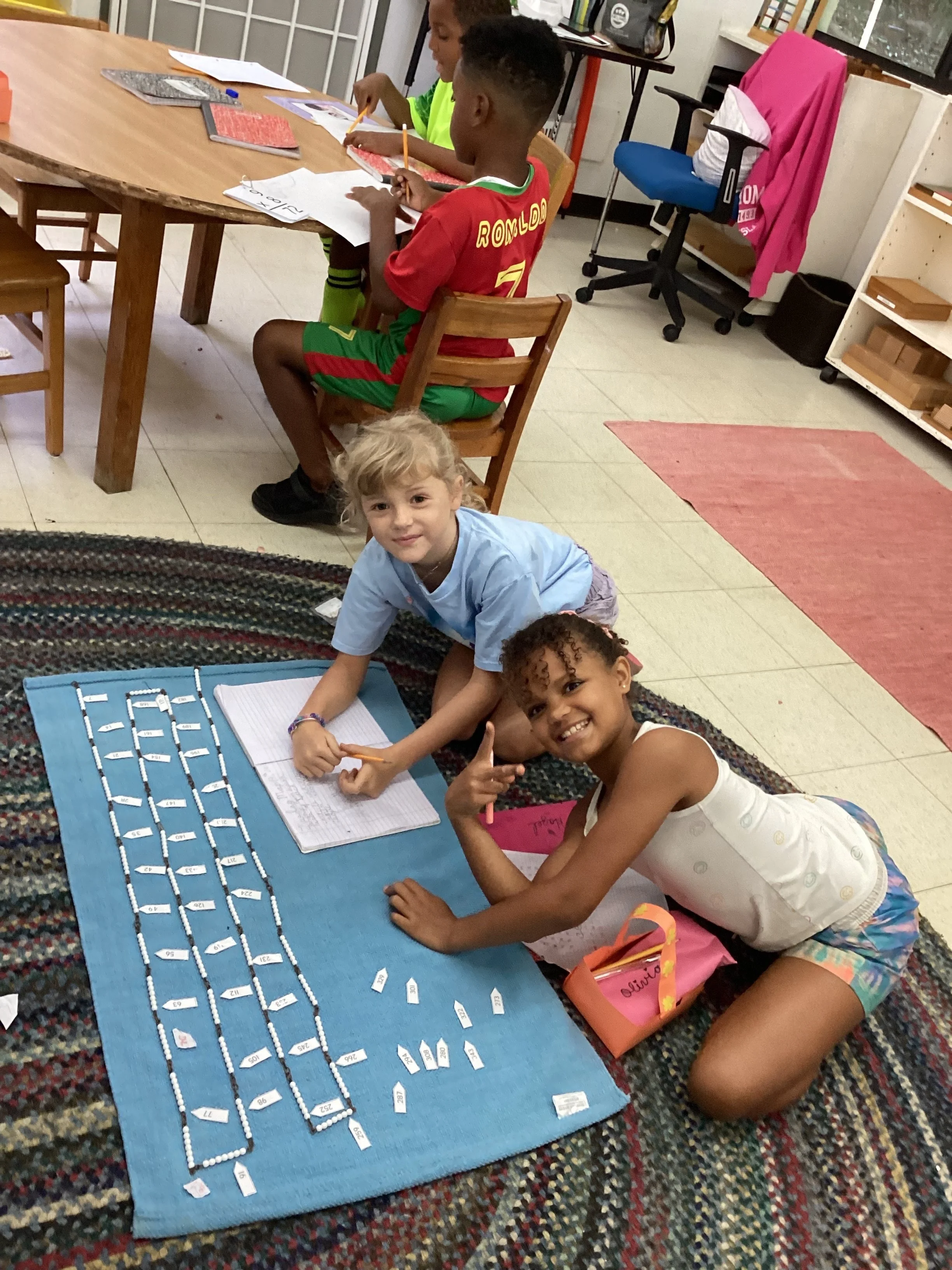 Three children engaging in a classroom activity with a large blue mat on the floor, exploring a number line made of small flags and a notebook, in a classroom setting.