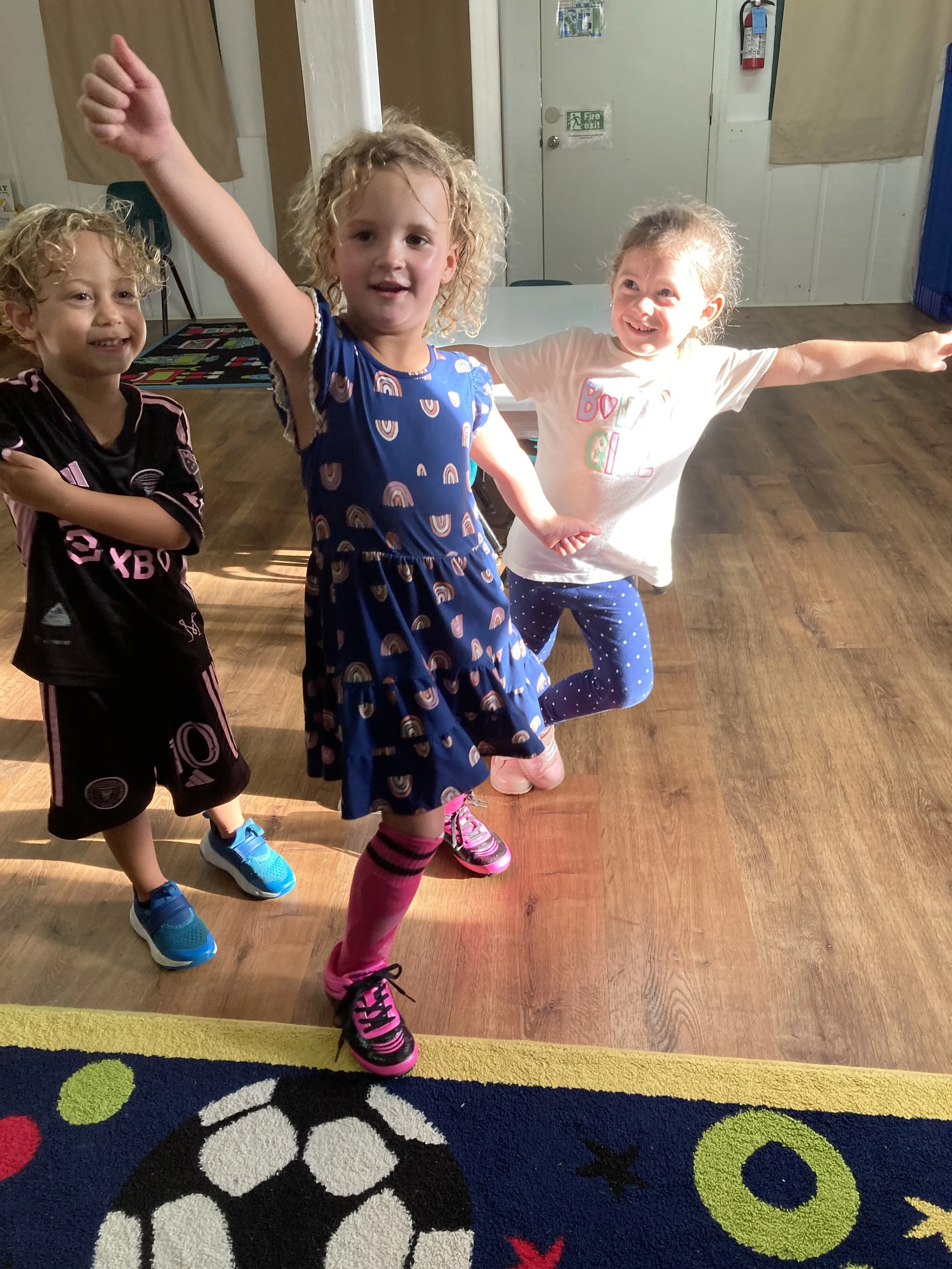 Three young girls dancing or playing together indoors, with one girl in a blue dress with rainbow patterns in front, wearing pink and black sneakers. The girl in the middle is wearing a white t-shirt with colorful text and dark blue leggings with white dots, while the girl on the left is in a black sports jersey and shorts with blue sneakers. All three are smiling and have their arms outstretched or crossed.
