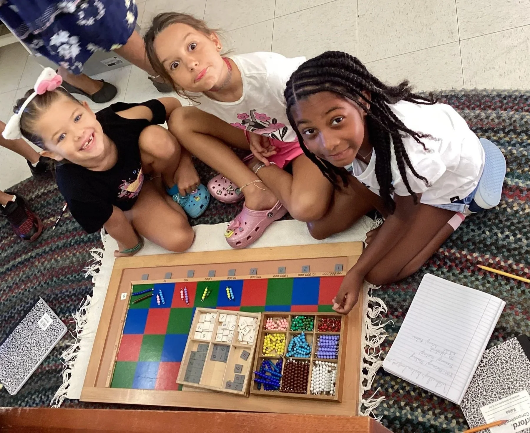 Three young girls sitting on a colorful rug around a wooden board game with colorful beads and tiles, looking up at the camera.