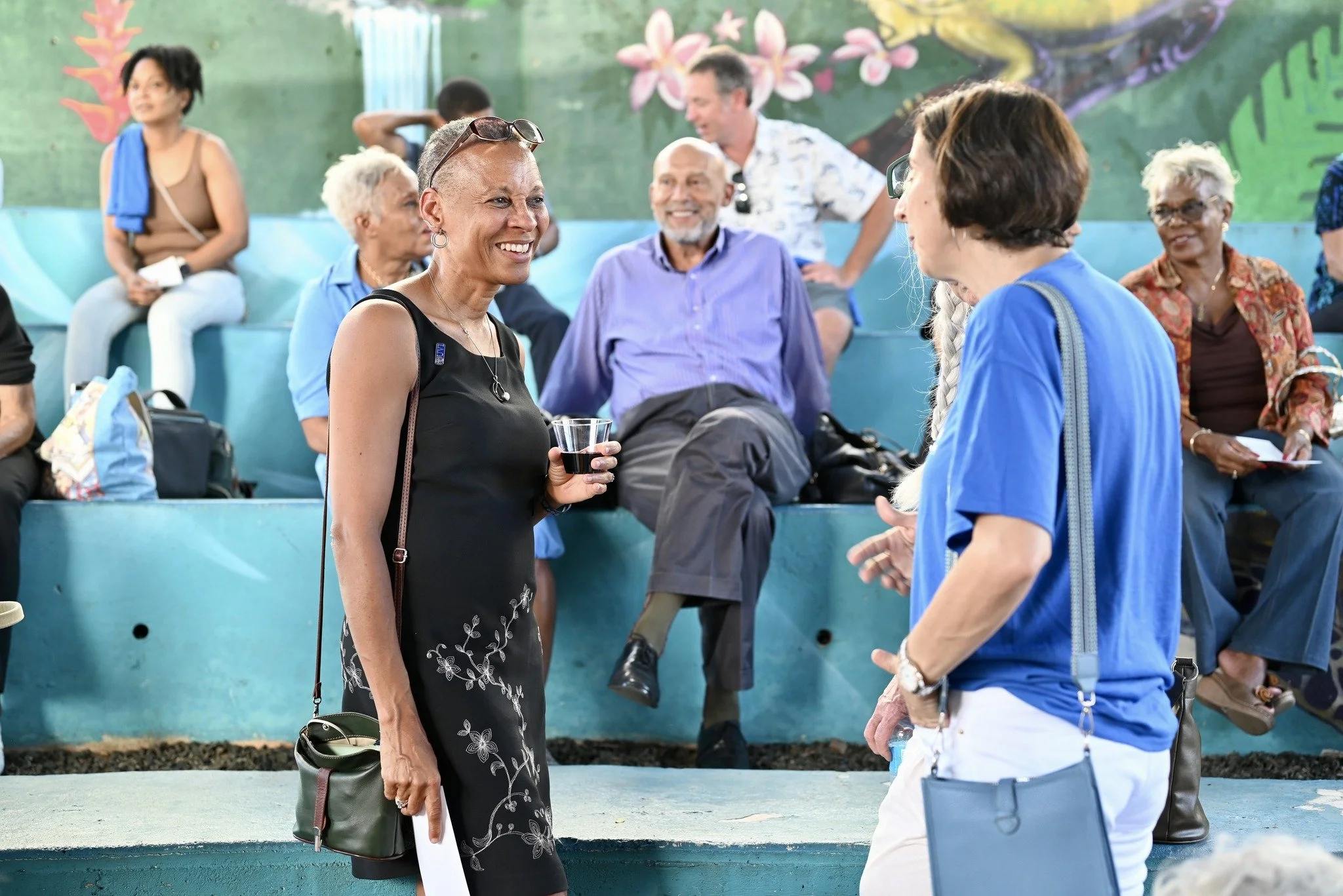 Two women are talking in the foreground, one holding a drink, smiling; they are surrounded by other seated and standing people in a colorful, mural-decorated setting.