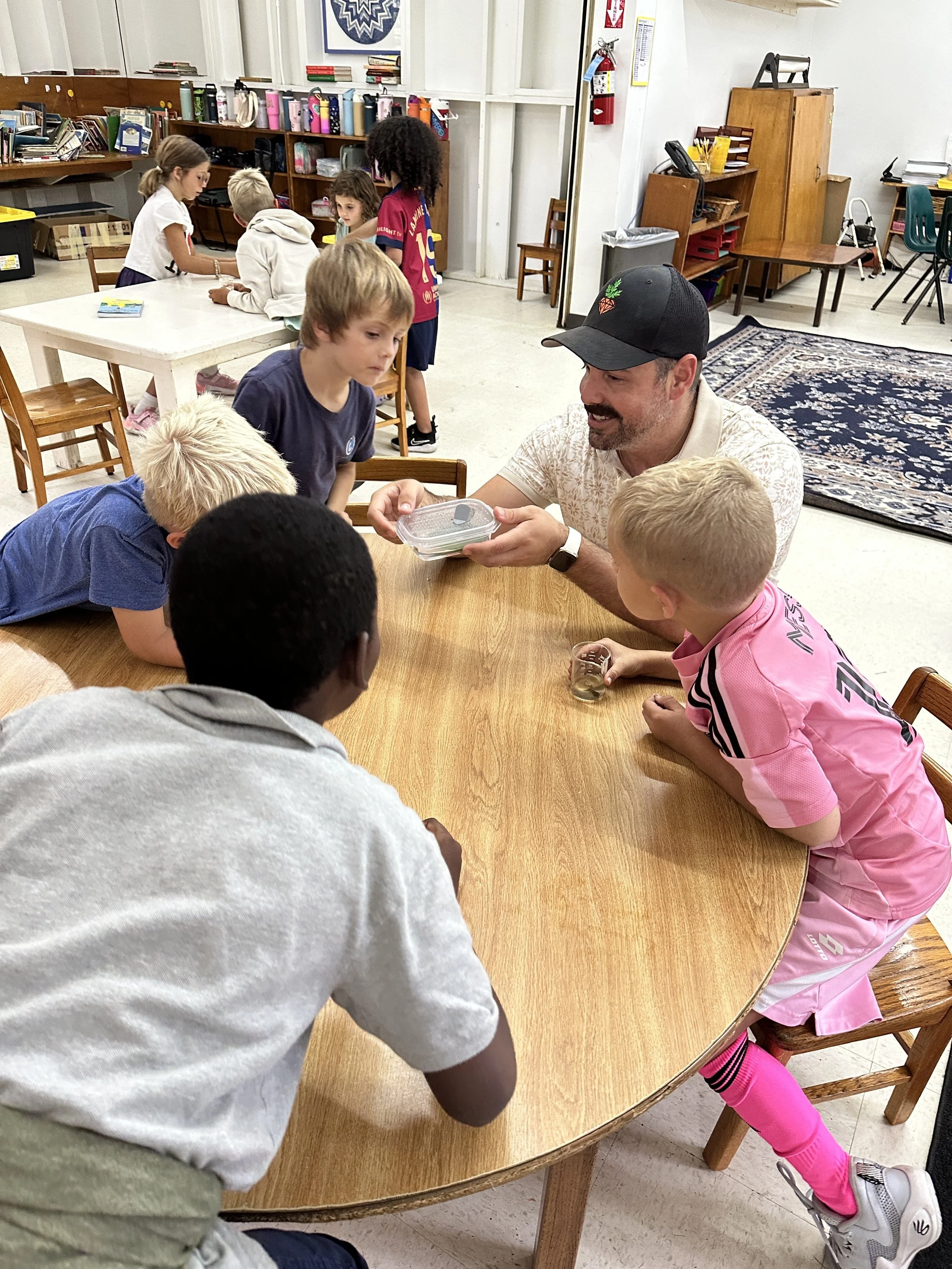 Teacher showing an insect to children sitting around a wooden table in a classroom.