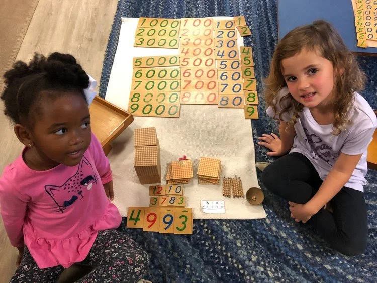 Two young girls sitting on a blue patterned rug with educational counting and multiplication materials, including number cards, small wooden blocks, and a dry erase board with numbers marked for a math activity.