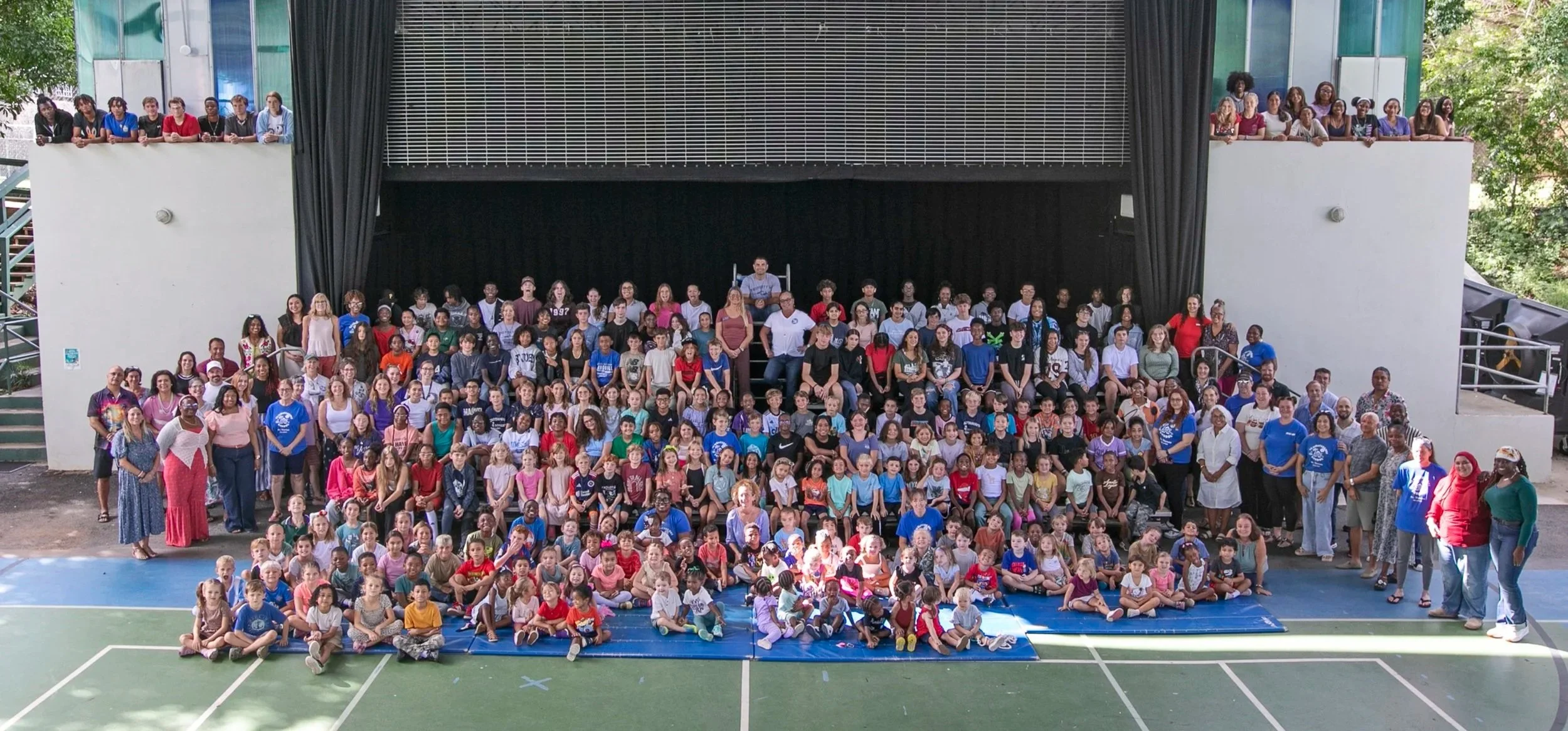 Large group of children and adults gathered in an outdoor gymnasium or sports court, posing for a group photo in front of a stage.