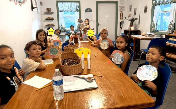 Children sitting around a long table in a classroom holding paper clocks they made, with craft supplies and water bottle on the table.