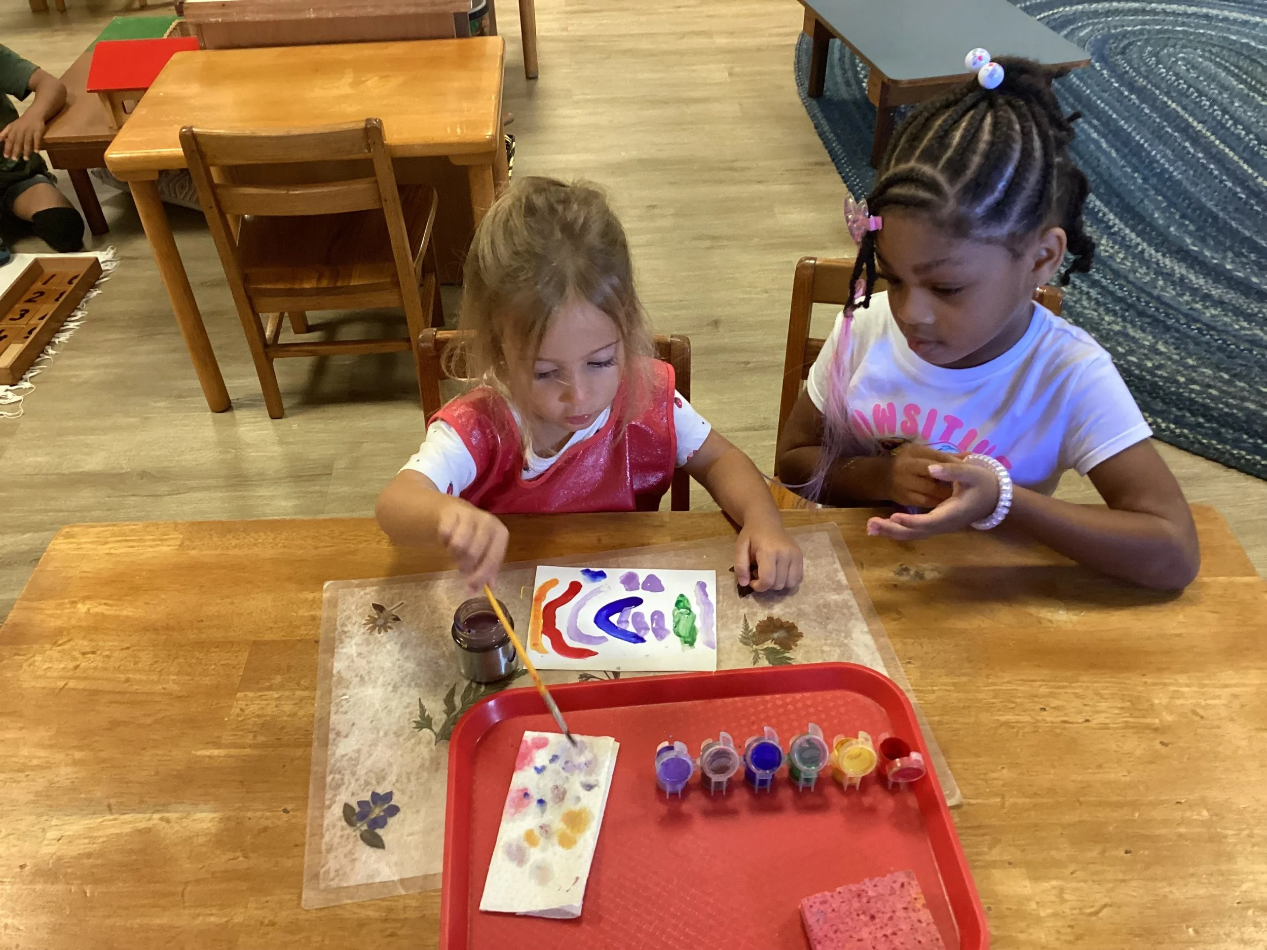 Two young girls sit at a wooden table, painting with bright colors on paper, with a tray of paint bottles and a paper towel in front of them.