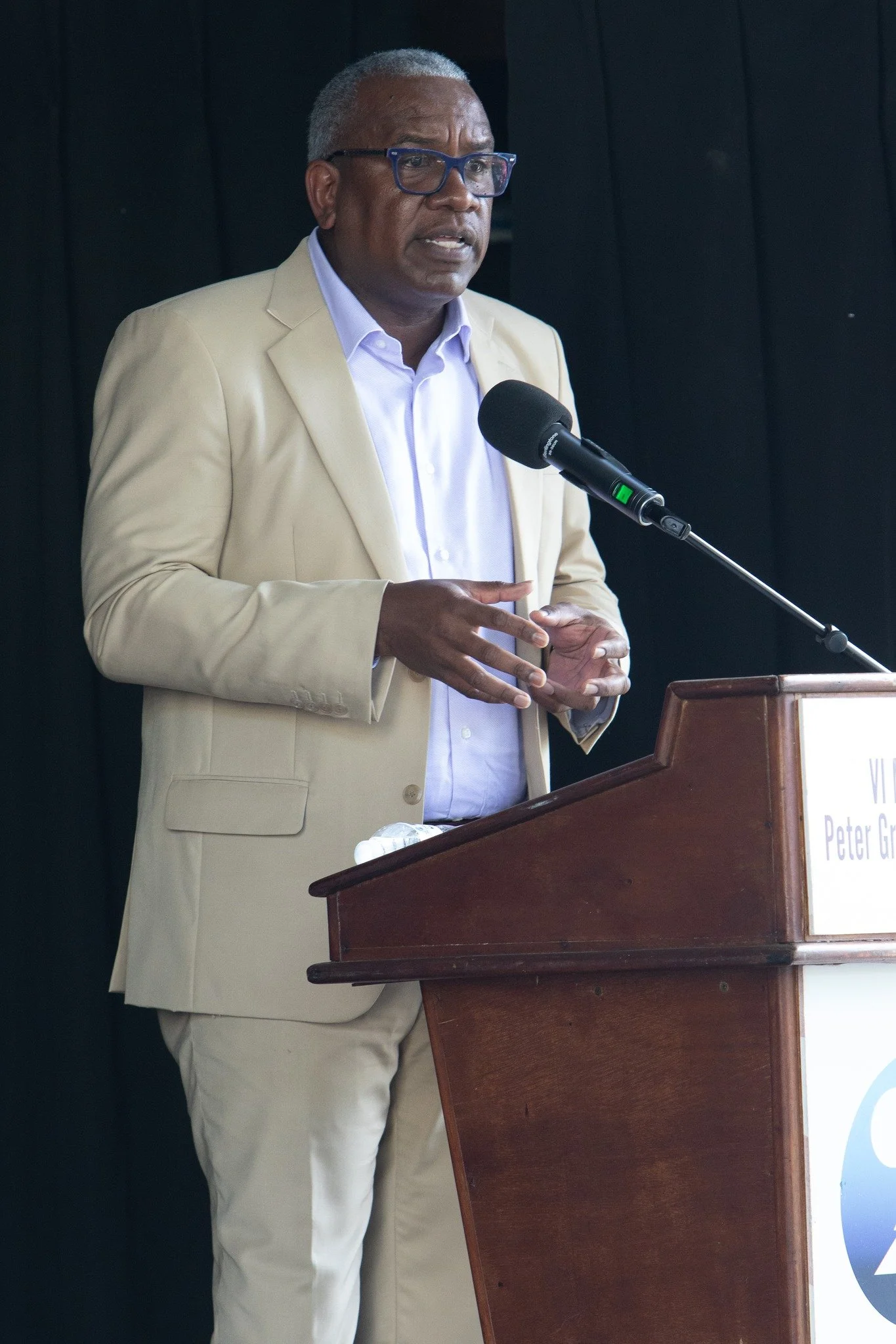 An African American man wearing glasses and a beige suit jacket speaking at a podium with a microphone.