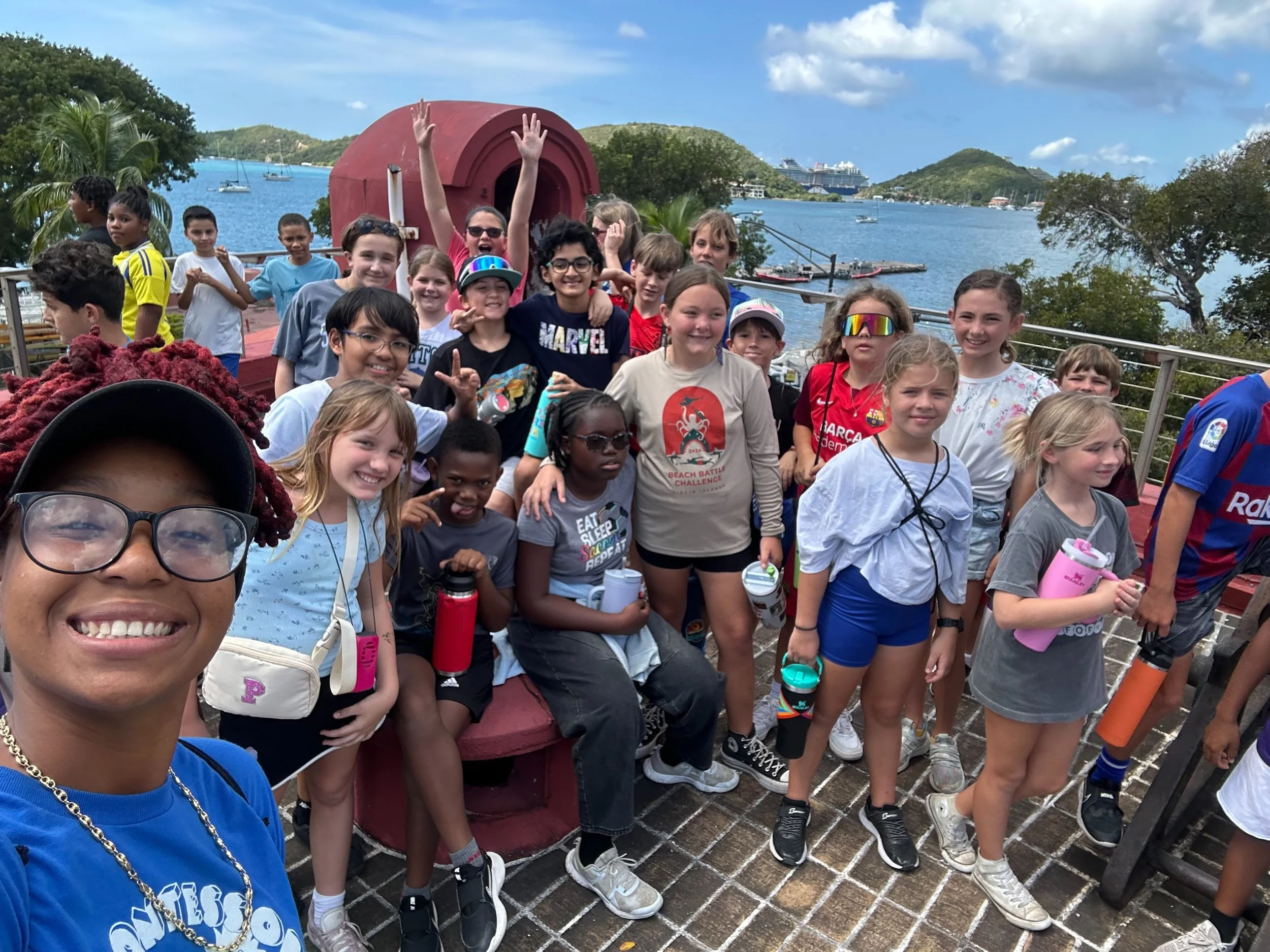 Group of children and a woman on a boat deck near water with lush green islands and cruise ships in the background.