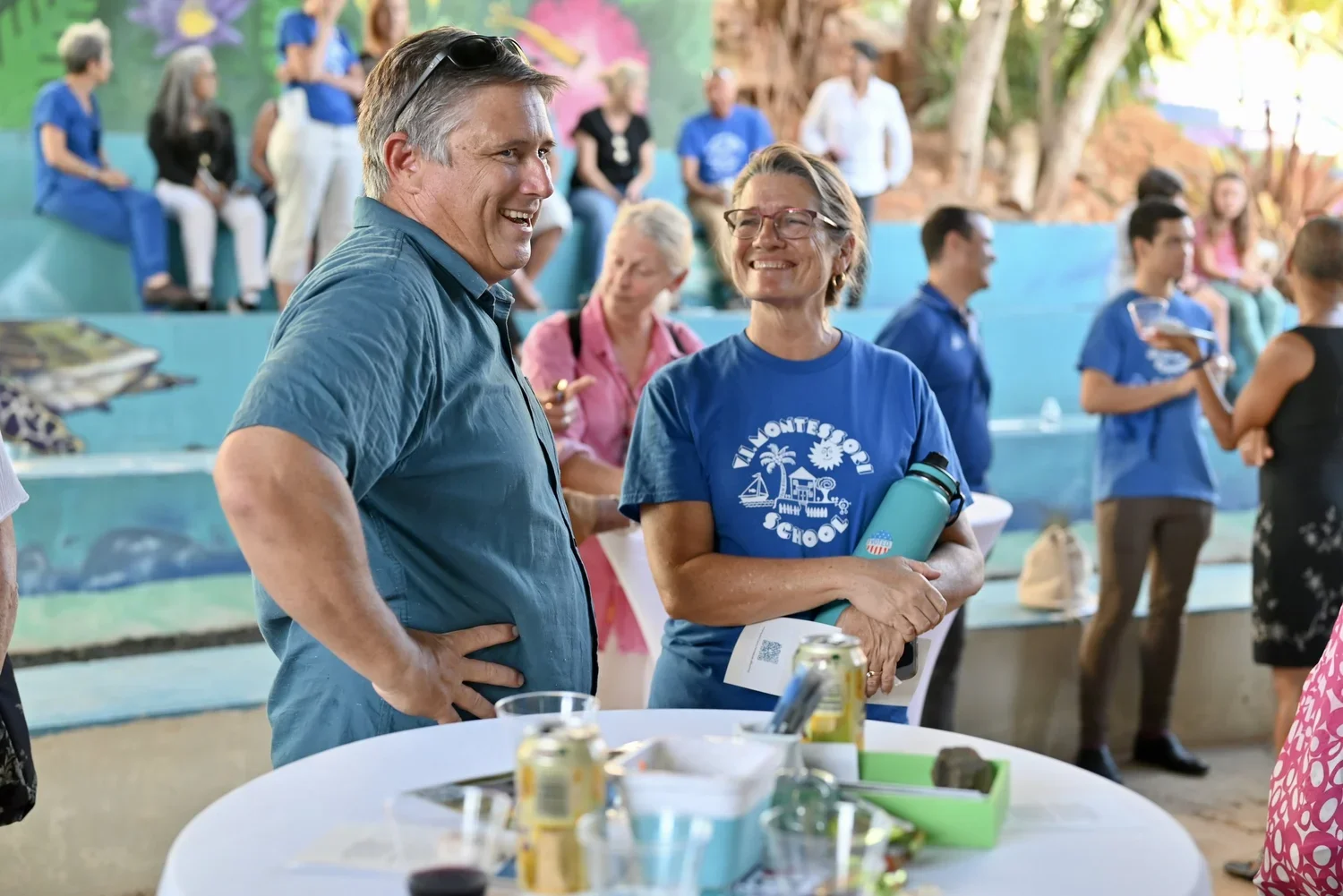 A group of people at an outdoor event, smiling and talking, with some seated and others standing, including a woman in a blue T-shirt with a school logo and a man in a blue shirt.