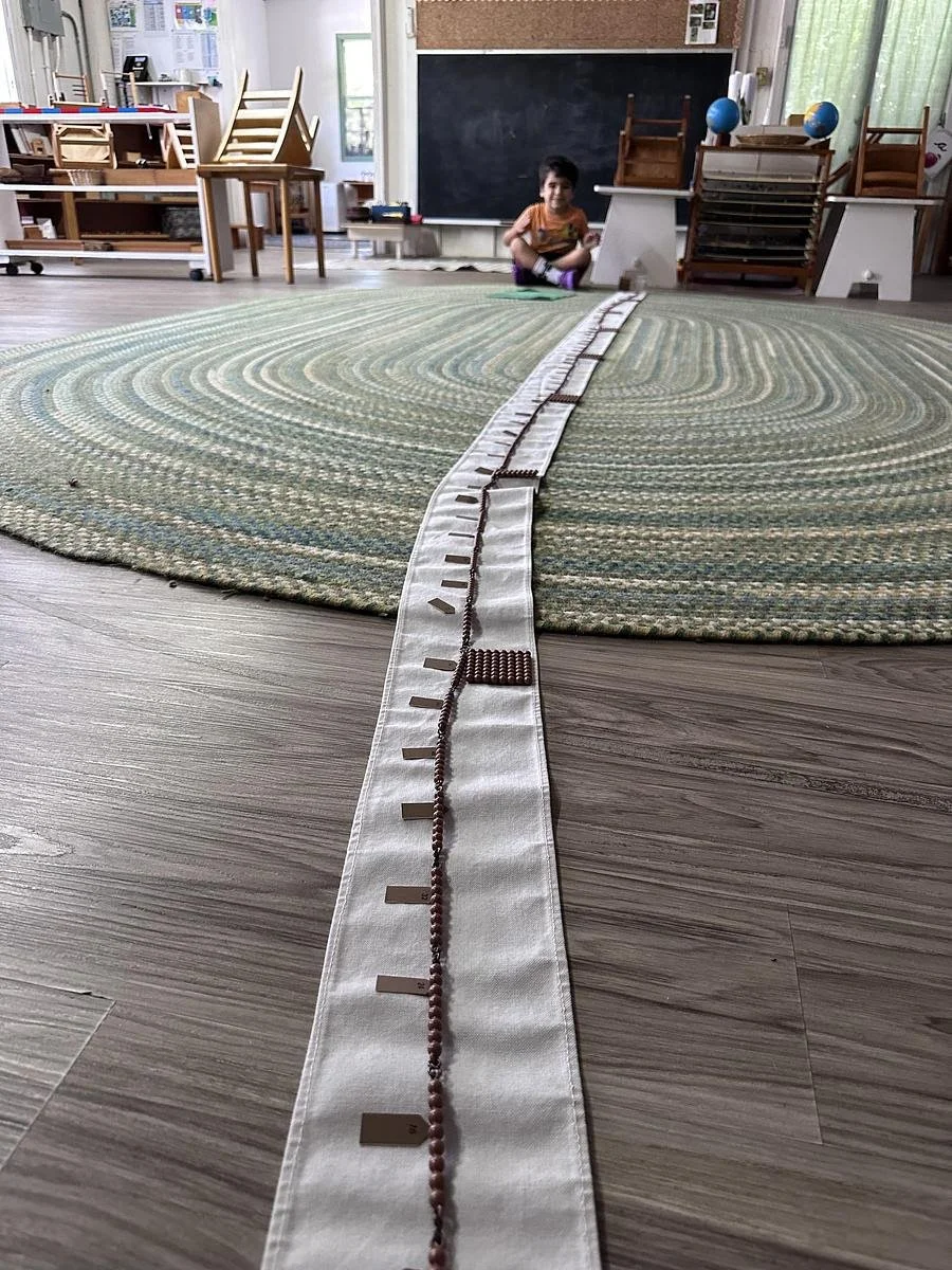 A child sitting on a green carpet in a classroom with chairs and educational materials, with a long strip of cloth and beads laid out on the floor in the foreground.