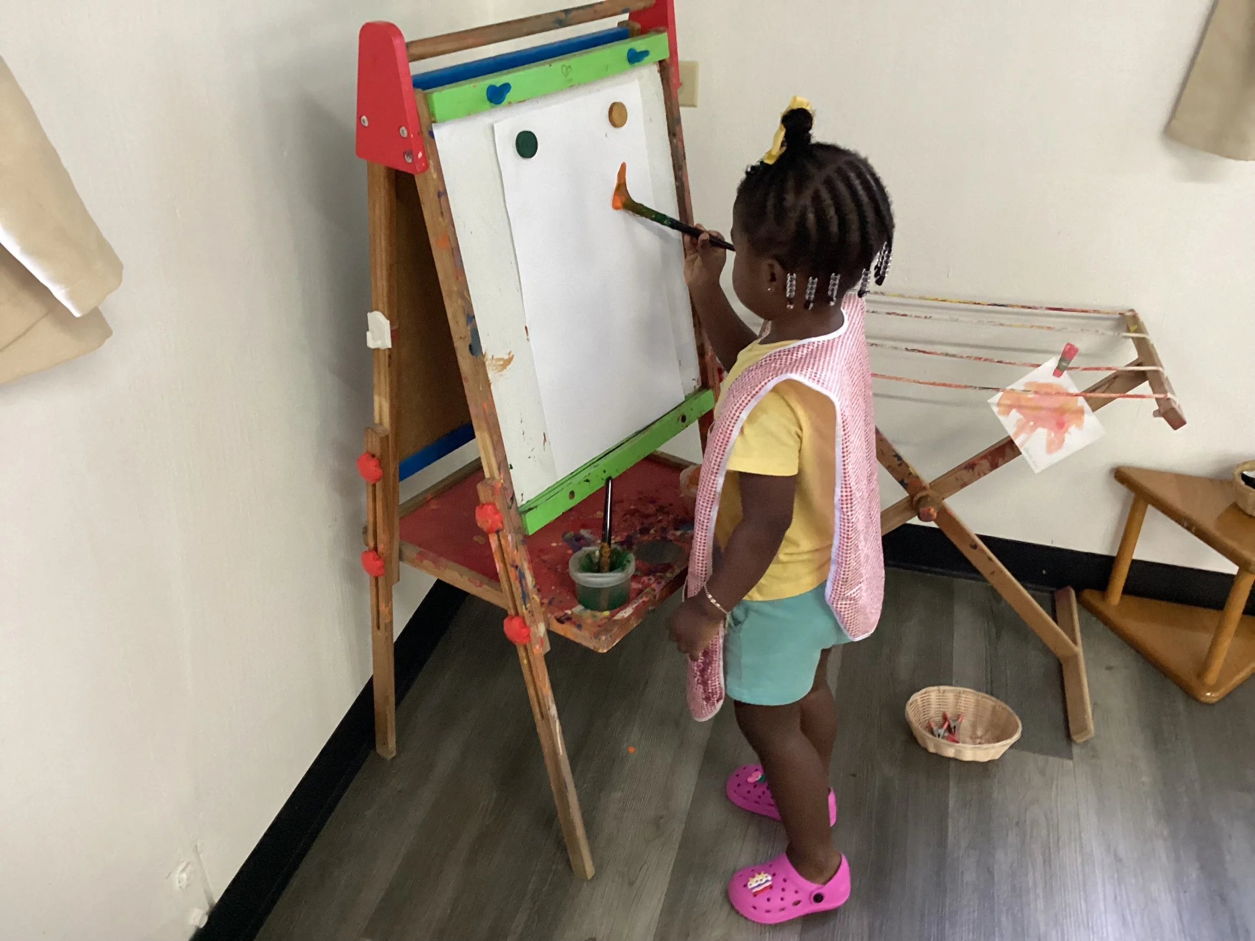 Young girl painting on a whiteboard easel in a room with wooden floors, pink Crocs, and a small basket of paint supplies nearby.