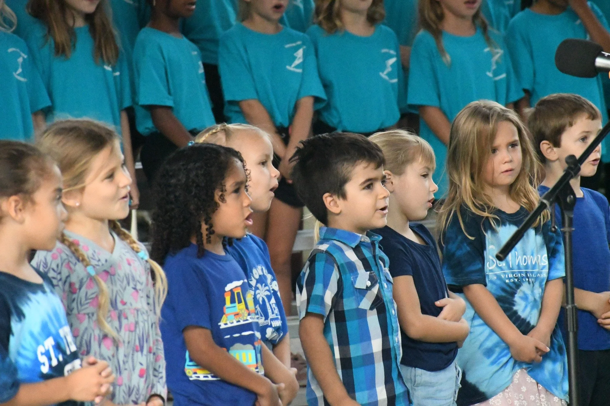 Children singing or performing in a choir, with some in front and others in the back, all wearing casual clothes, in a school or community event setting.