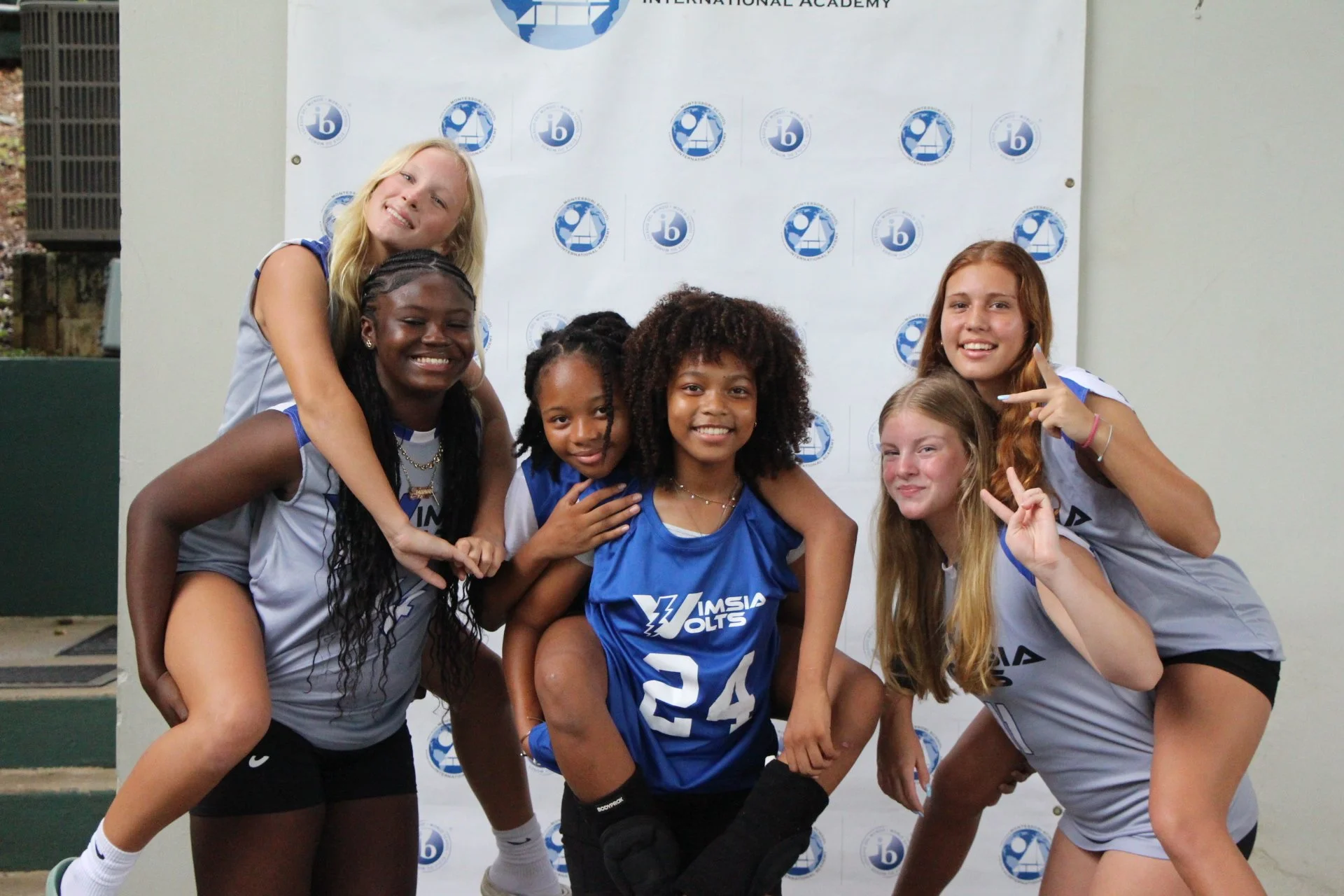 Group of young female volleyball players in sports uniforms posing together, smiling and making peace signs, in front of a white backdrop with logos.
