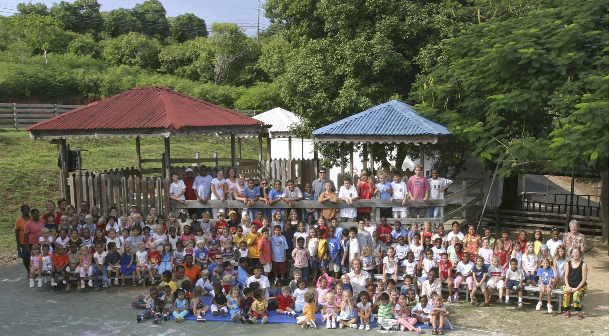 Group photo of children and adults outdoors in front of a playground structure with colorful roofs surrounded by green trees, during daytime.