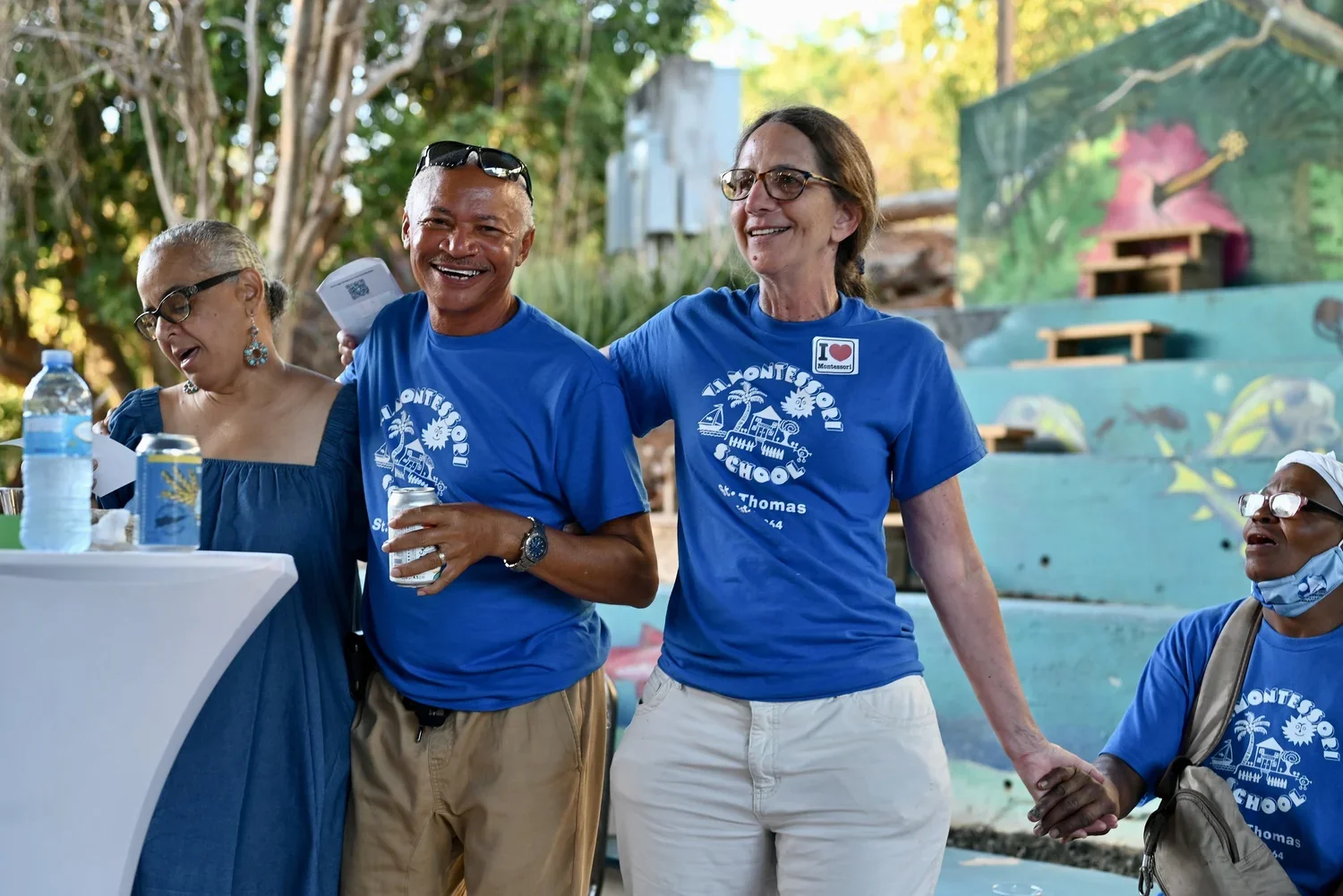 A group of diverse people smiling, holding hands, and wearing blue T-shirts with 'Almonte School' printed on them, standing outdoors at a school event or gathering.