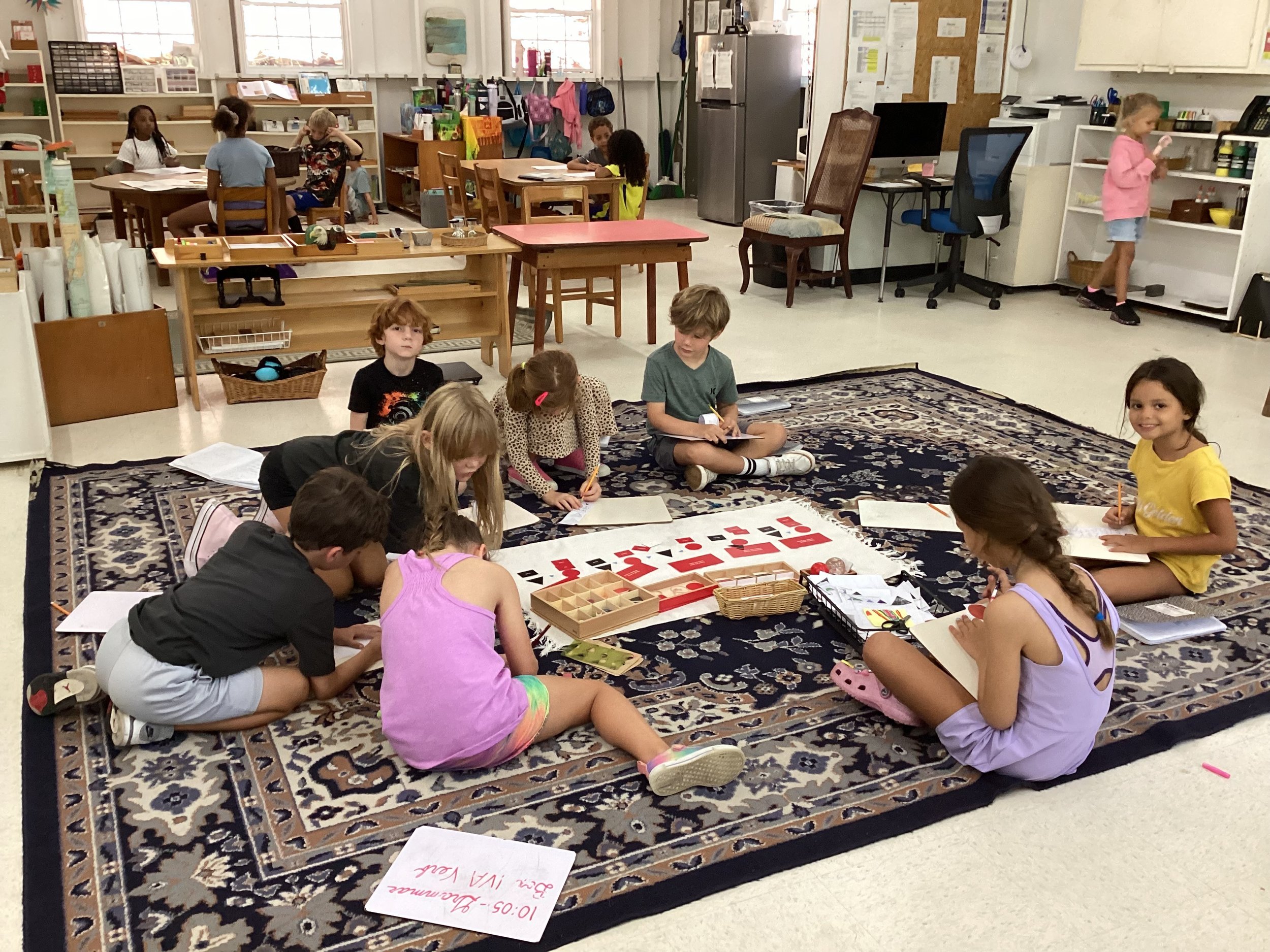 Group of young children sitting on a rug in a classroom, working on activities with papers and red and black tiles, with classroom furniture and supplies visible in the background.