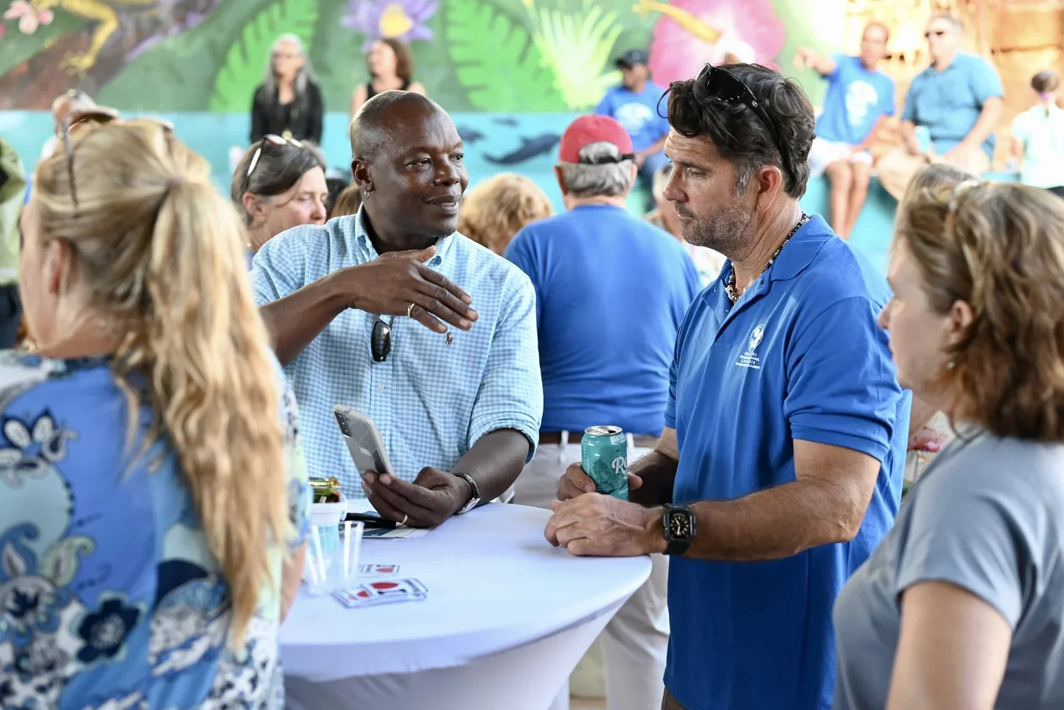 A diverse group of people gathered at a social event, engaging in conversation, with some holding drinks. The background features a mural of a vibrant, tropical scene.