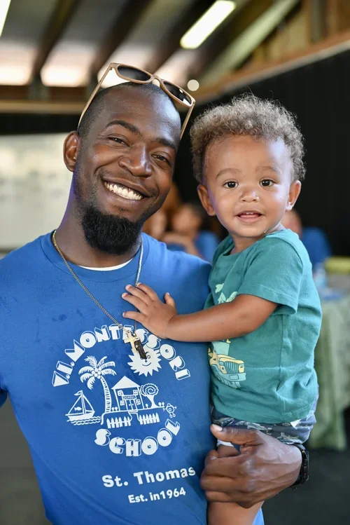 A man smiling while holding a small child in his arms. They are both looking at the camera. The man is wearing a blue T-shirt and sunglasses, and the child is wearing a green T-shirt.