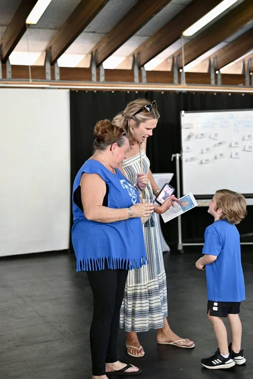 Two women and a young boy standing in a room with wooden ceiling beams, engaging in conversation using a smartphone and a notepad, with a whiteboard in the background.