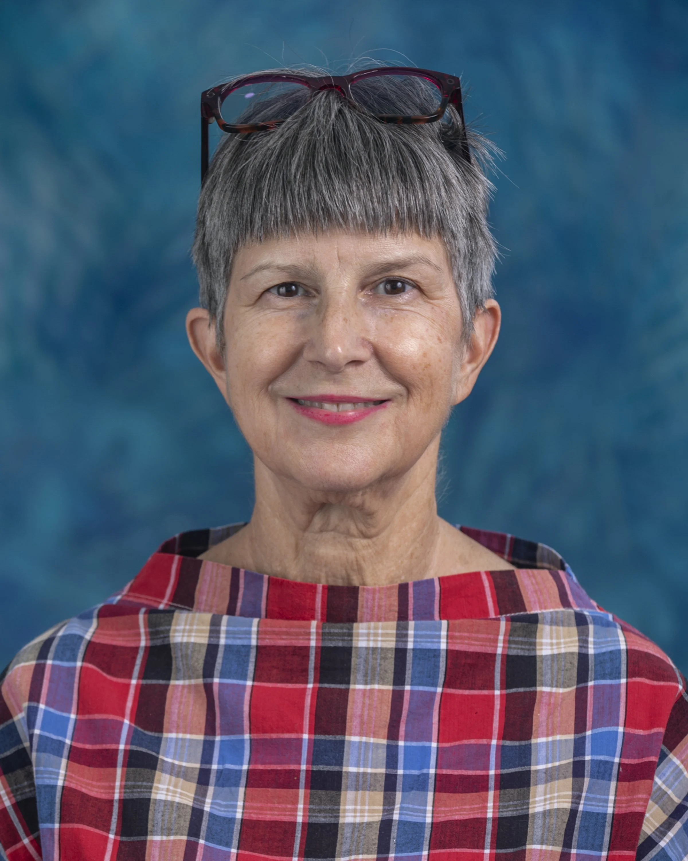 A woman with short styled gray hair and wearing large glasses, smiling for a selfie inside a store or café with wooden shelves displaying pastel-colored teapots and cups.
