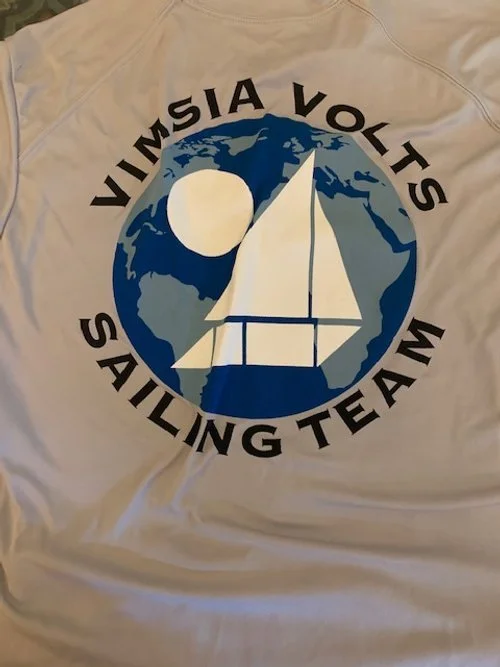 A logo on a fabric background featuring a blue and gray Earth with a white sailboat and a white circle, surrounded by the text 'VIMSIA VOY SAILING TEAM' in black.