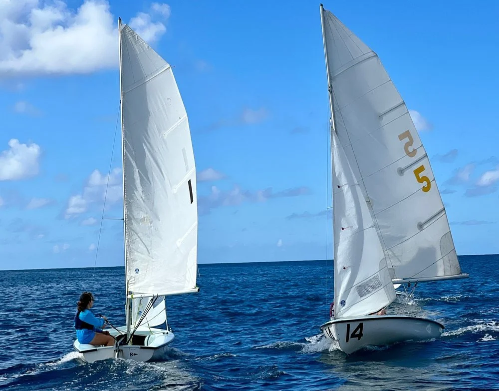 Two sailboats with white sails racing on the ocean under a partly cloudy sky.