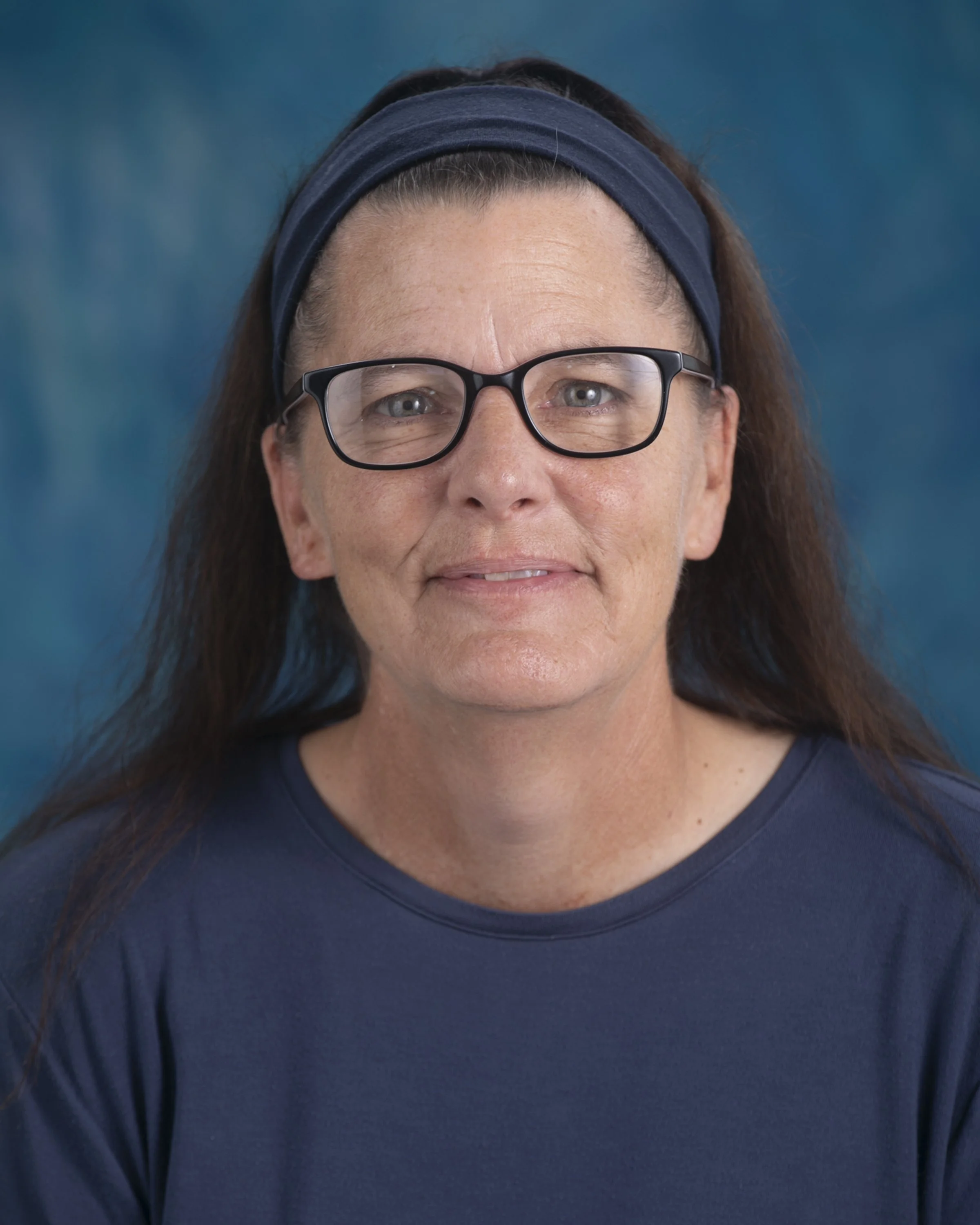 A woman with glasses and long dark hair, smiling, wearing a blue shirt and headband, standing outdoors with tropical plants and a wooden fence in the background.
