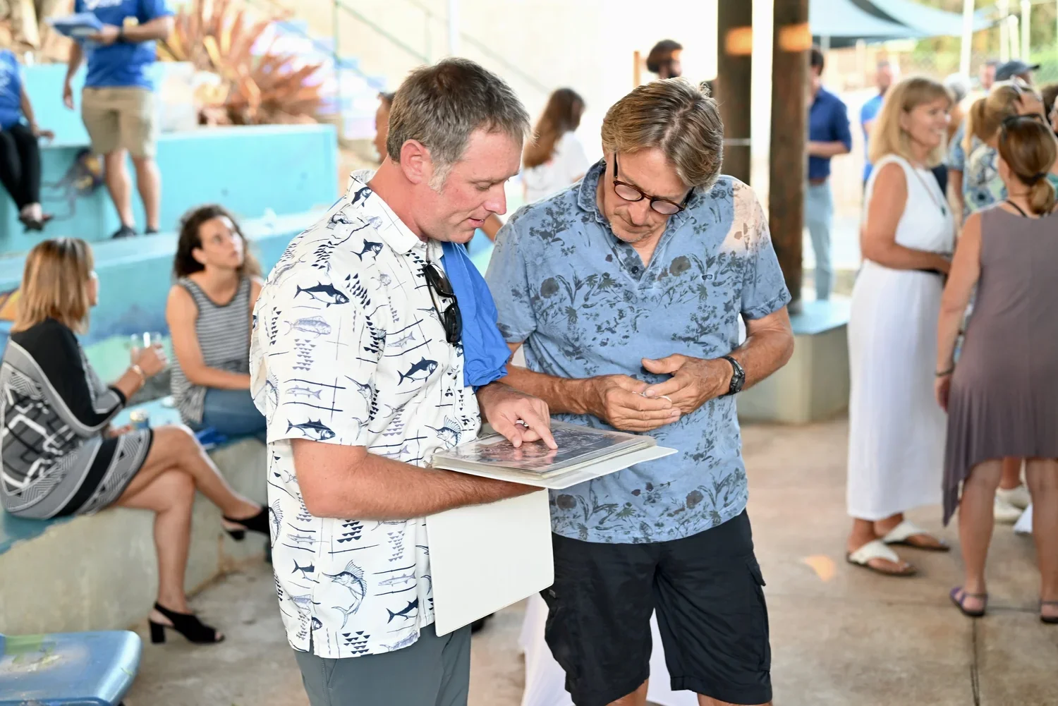 Two men are looking at a photo album at a social gathering, with other people talking and sitting in the background.