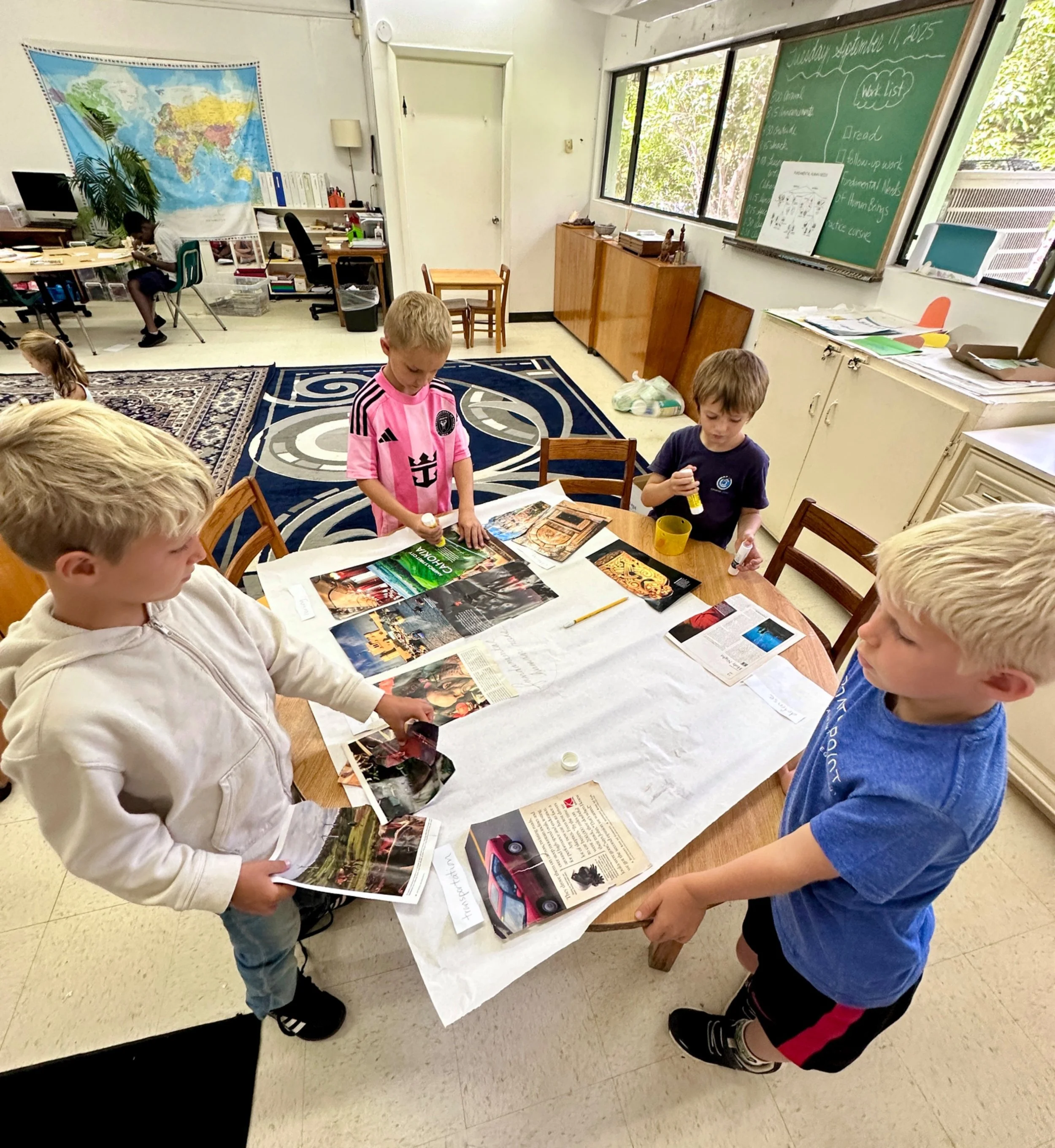 Four children working on a collage project in a classroom, with magazines and scissors on a table.