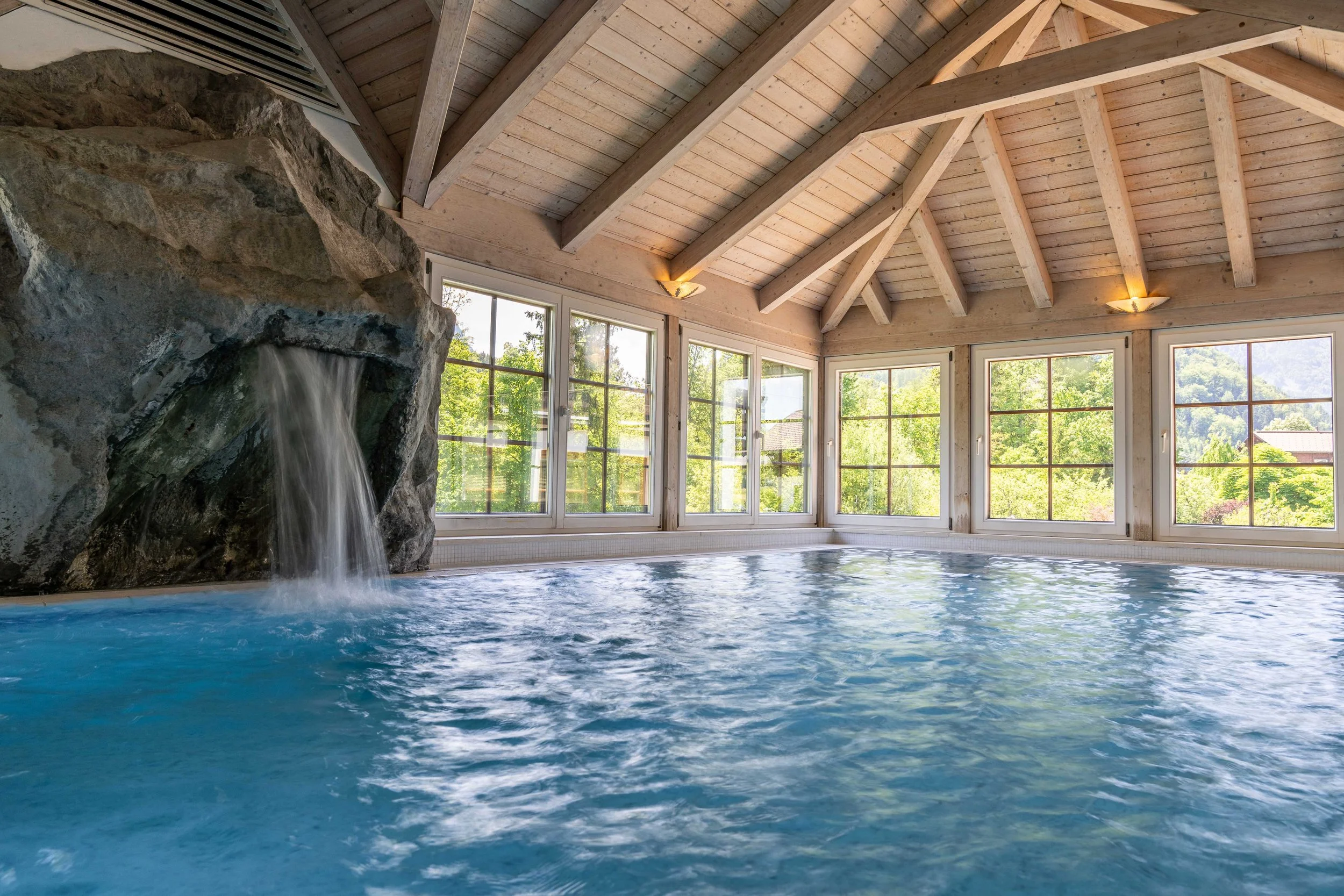 Innenaufnahme eines beheizten Schwimmbeckens mit Wasserfall, große Fenster mit Blick in die Natur