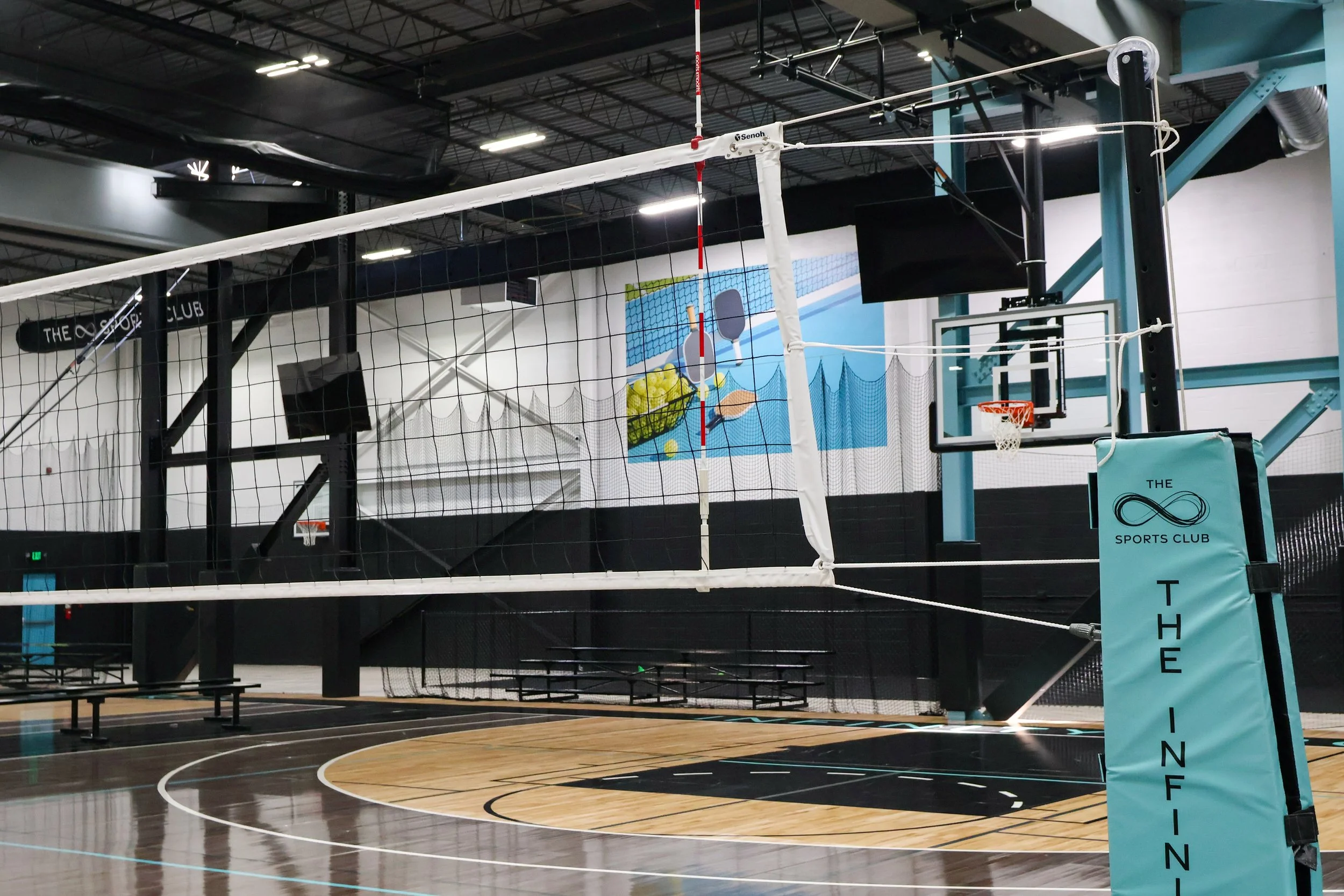 Indoor sports court with volleyball net, basketball hoop, and a volleyball logo mural on the wall, in a gym with high ceilings and bright overhead lighting.