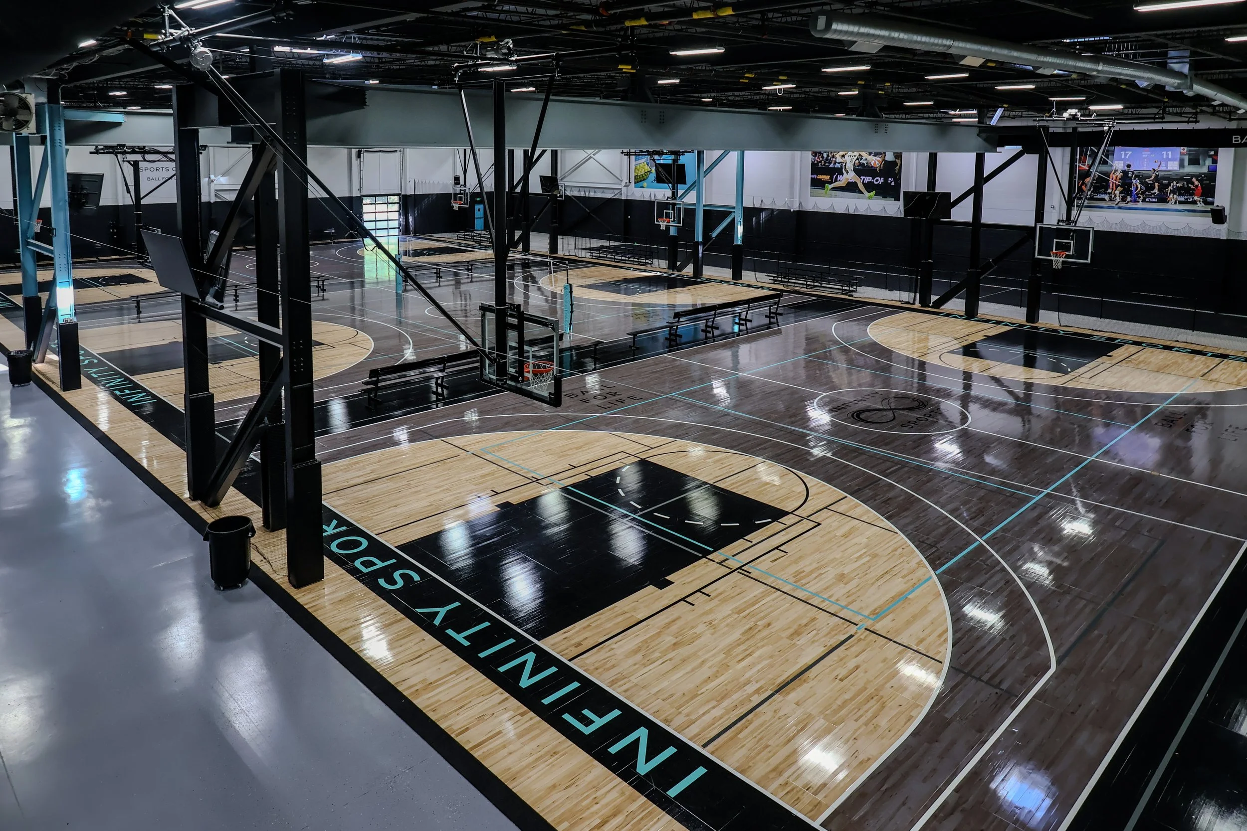 Indoor basketball court with multiple hoops, black and natural wood flooring, benches, digital screens, and overhead lighting.