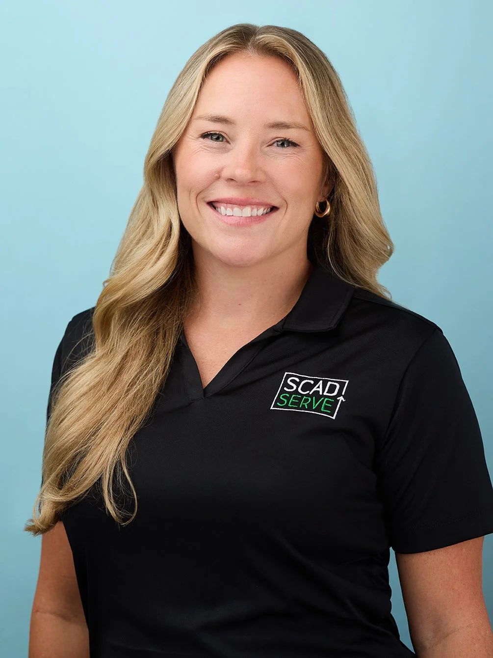 A woman with long blonde hair, wearing a black polo shirt with a logo that says 'SCAD Serve,' smiling, standing against a blue background.