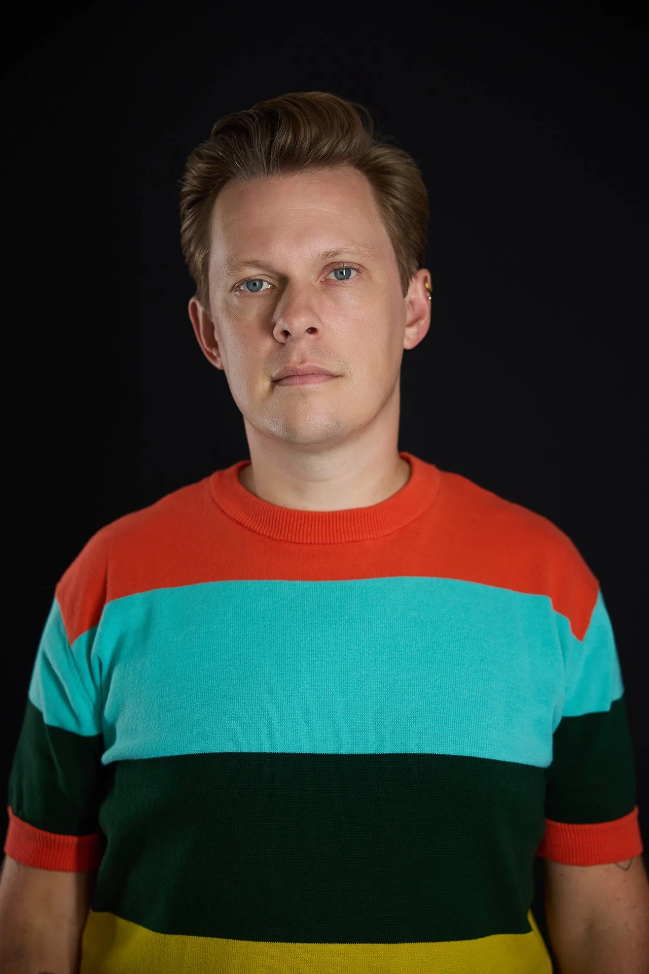 Portrait of a young man with blonde hair and blue eyes wearing a multicolored striped T-shirt against a black background.