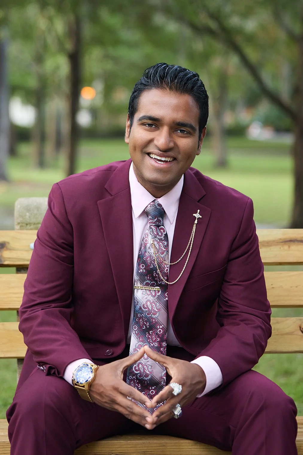 A smiling man dressed in a maroon suit, white shirt, patterned tie, and accessorized with jewelry, sitting on a wooden bench outdoors in a park with trees in the background.