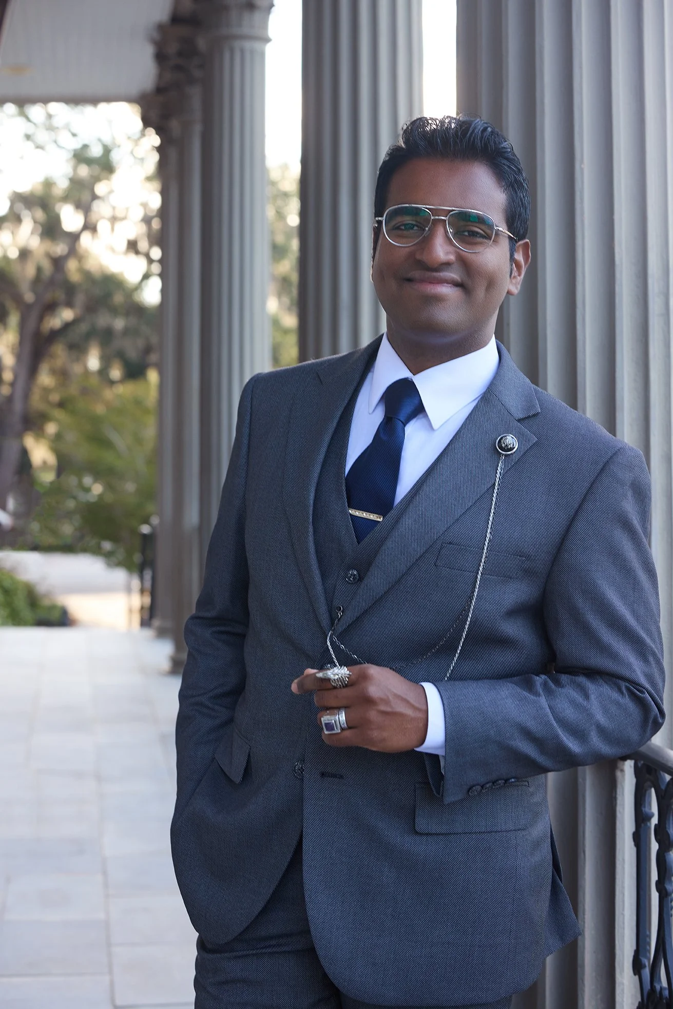 A man in a gray suit, white shirt, and navy tie standing outdoors on a stone pathway with classical columns in the background. He is holding a pocket watch chain and wearing glasses.
