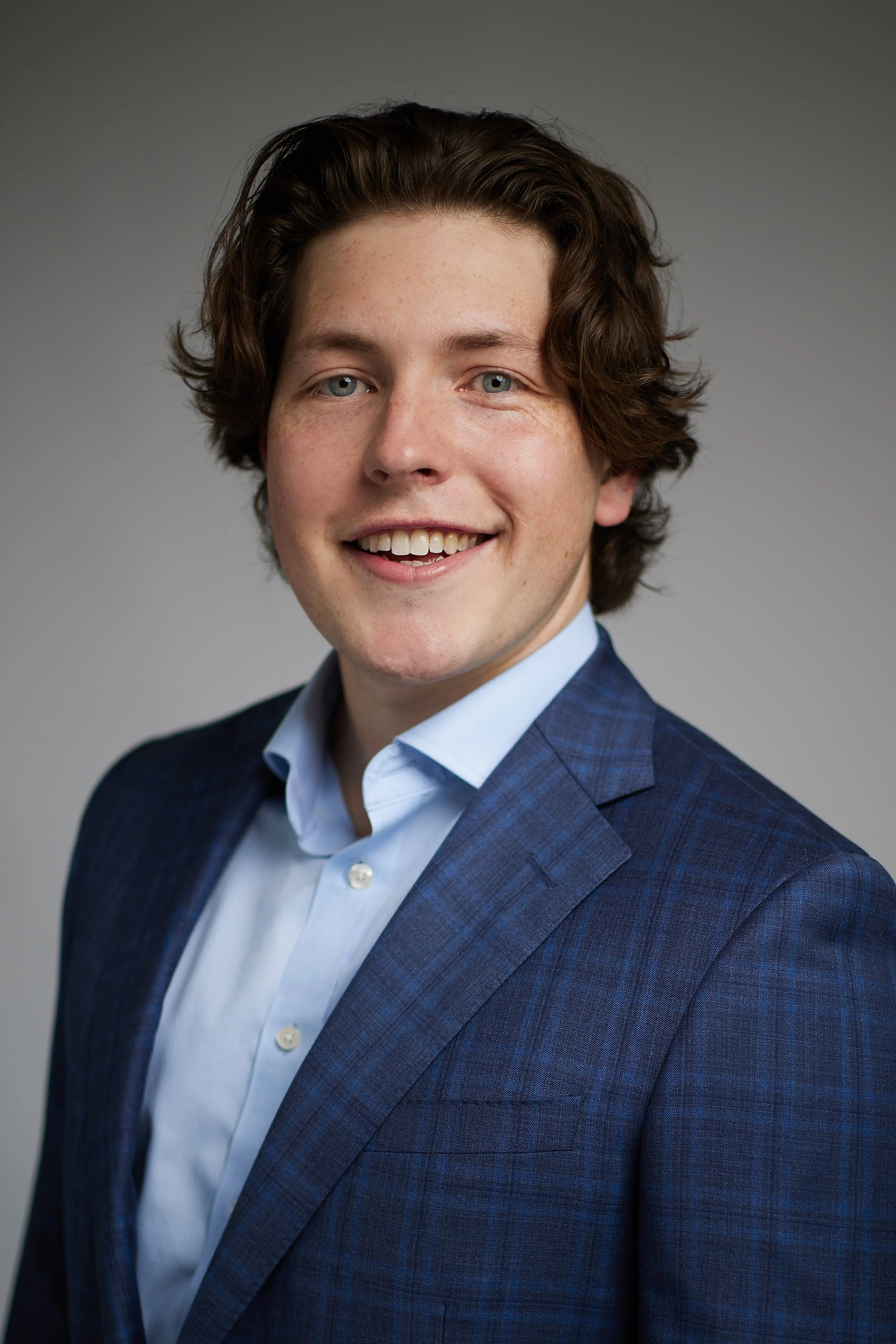 Headshot of a young man with wavy brown hair, blue eyes, and fair skin, wearing a blue plaid suit jacket and white shirt, smiling against a gray background.