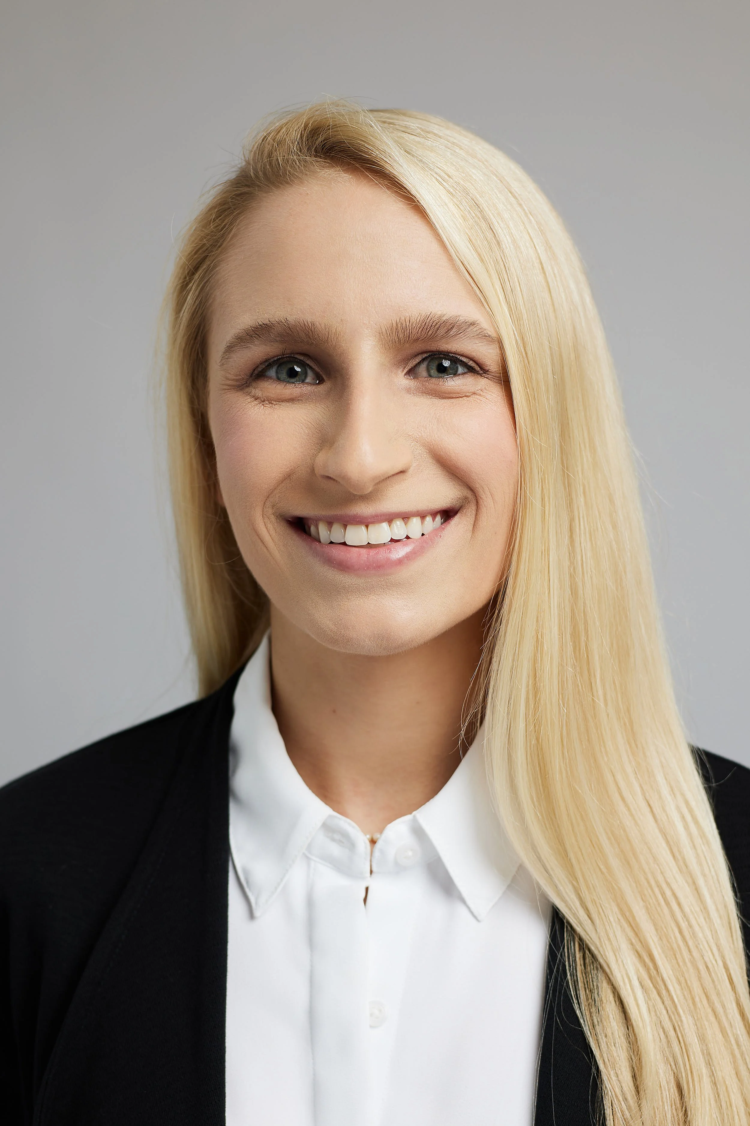 A professional woman with long blonde hair, wearing a black blazer and white shirt, smiling at the camera.
