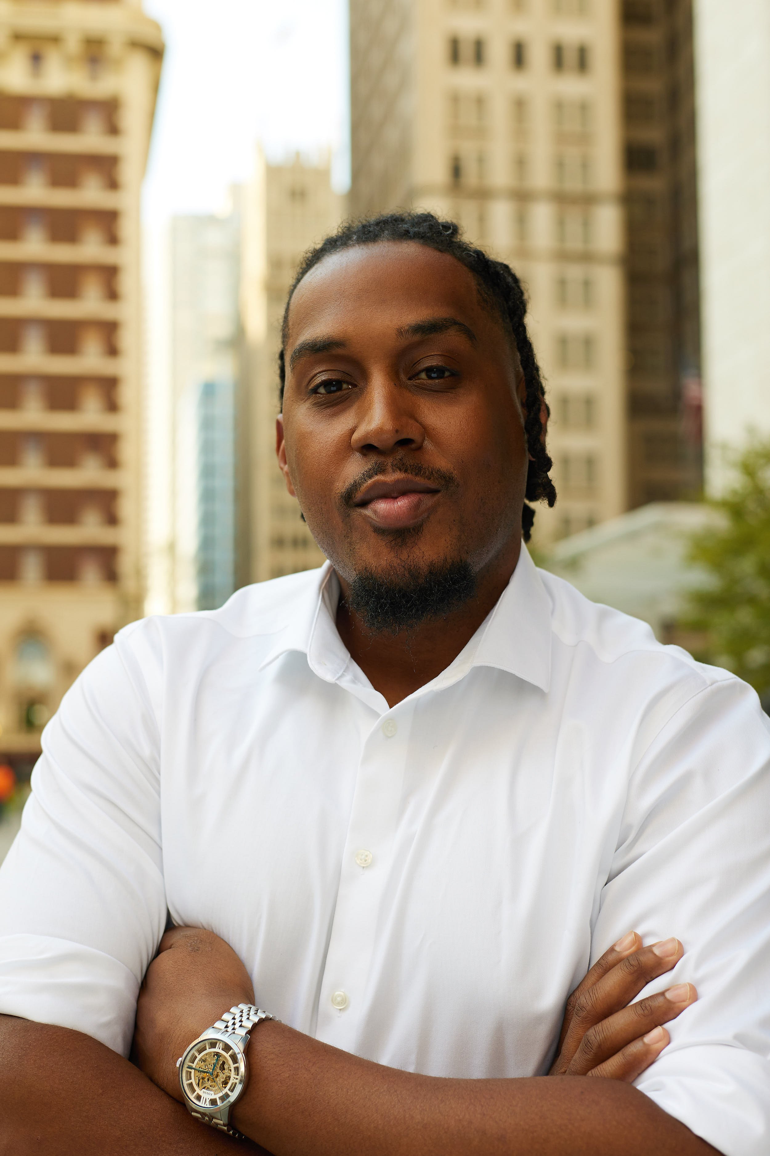 A man with dreadlocks and a goatee, wearing a white dress shirt and a silver watch, standing outdoors in an urban setting surrounded by tall buildings.