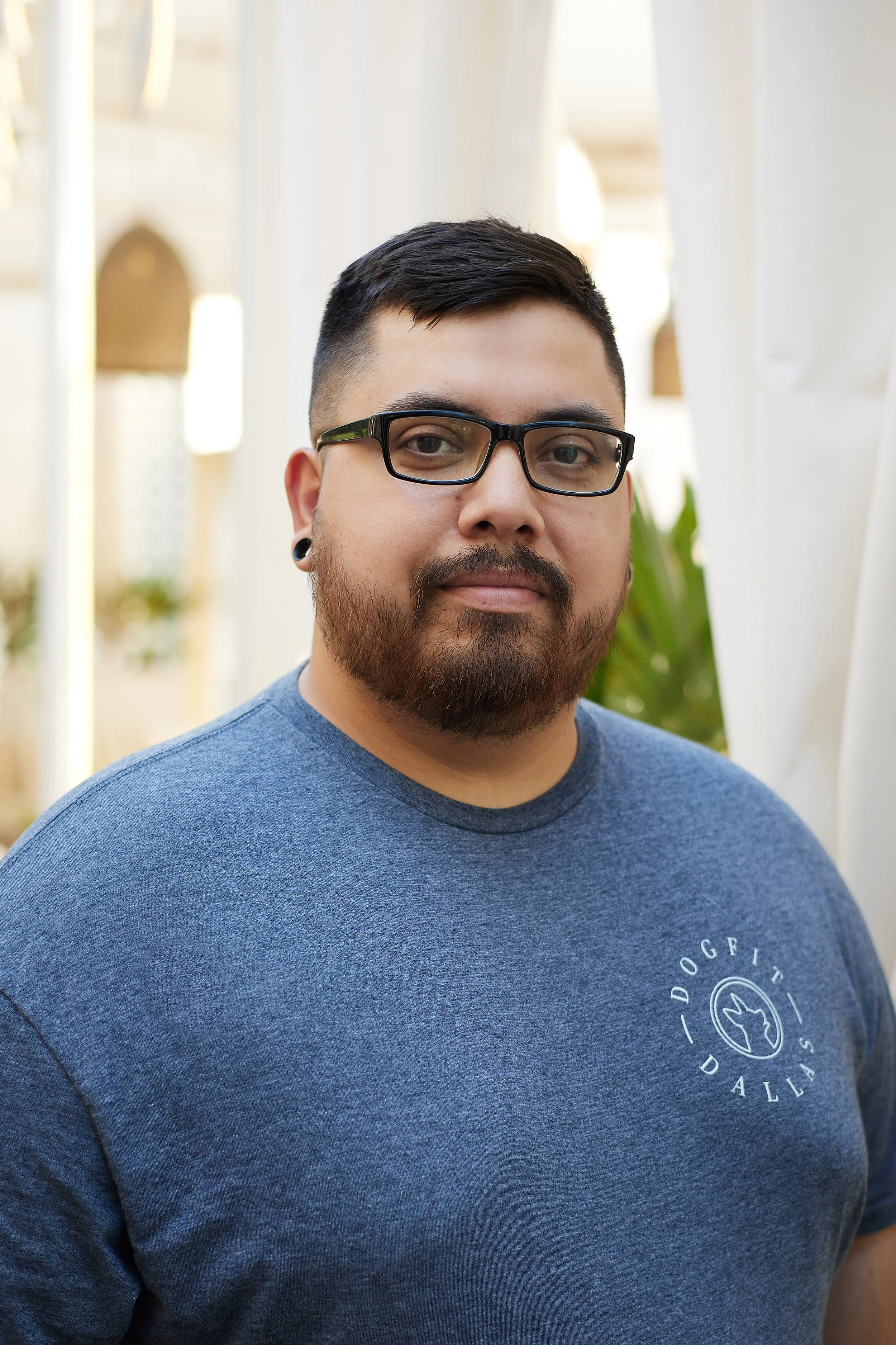 A man with glasses and a beard wearing a blue t-shirt with a dog logo, standing outdoors.