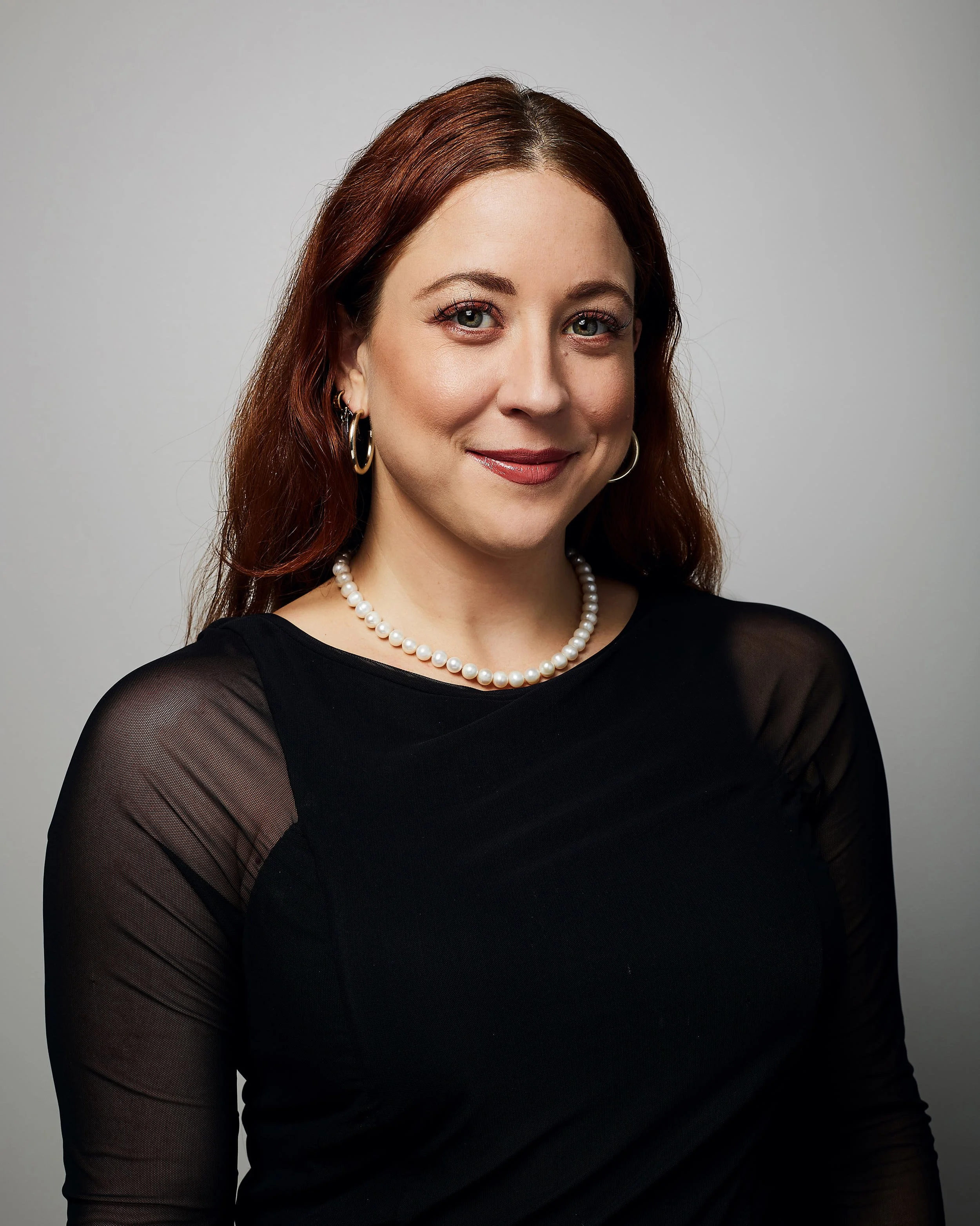 A woman with long, auburn hair and fair skin, wearing a black top with sheer sleeves, a pearl necklace, and gold hoop earrings, smiling against a plain background.